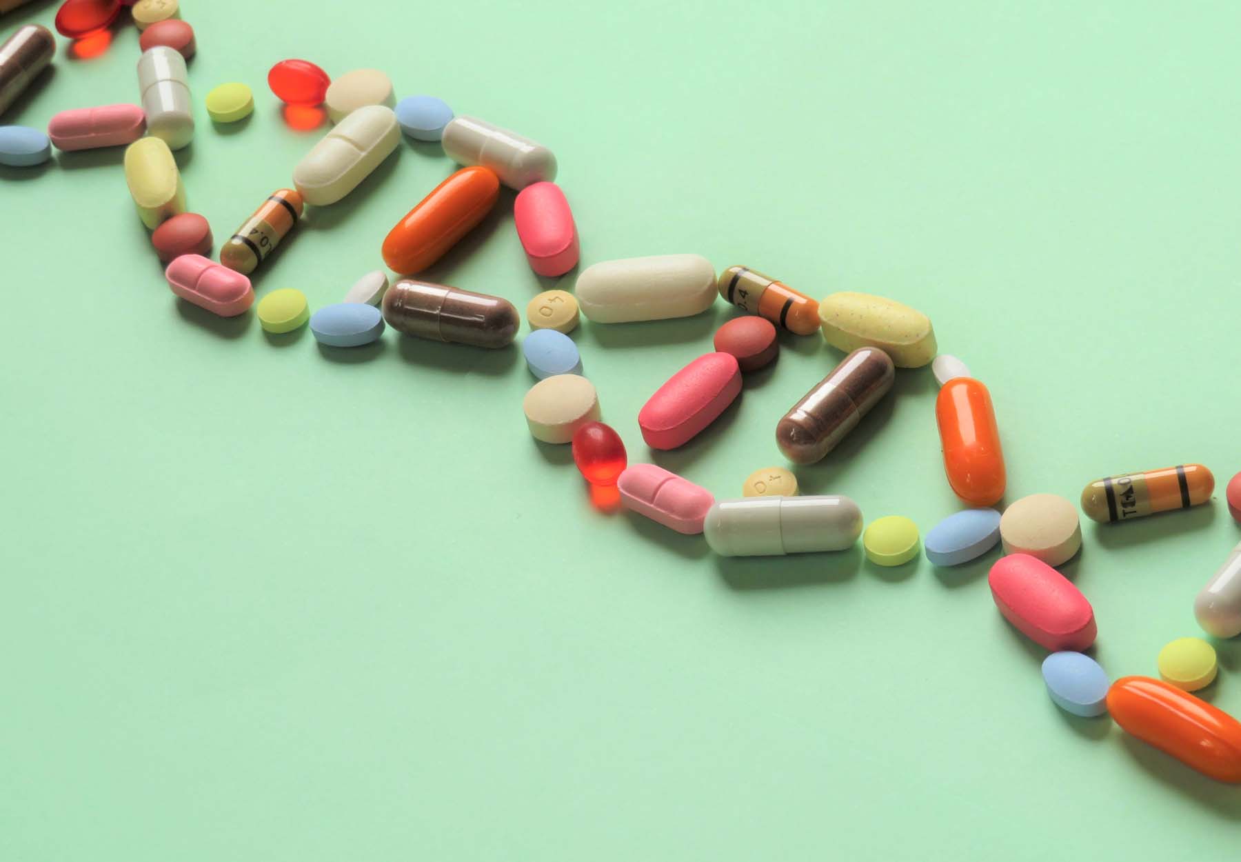 Horizontal image of prescription capsules and tablets of various shapes and colors arranged diagonally in the shape of DNA to represent pharmacogenetics. Green background.
