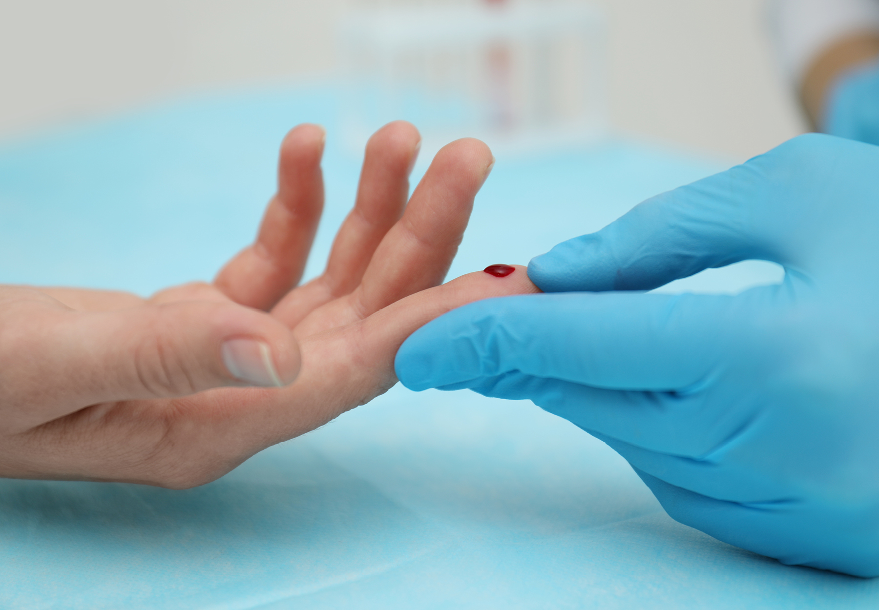 A doctor or nurse's blue-gloved hand holds a patient's hand that has just had a fingerstick test done. There is a drop of blood on the patient's index finger.