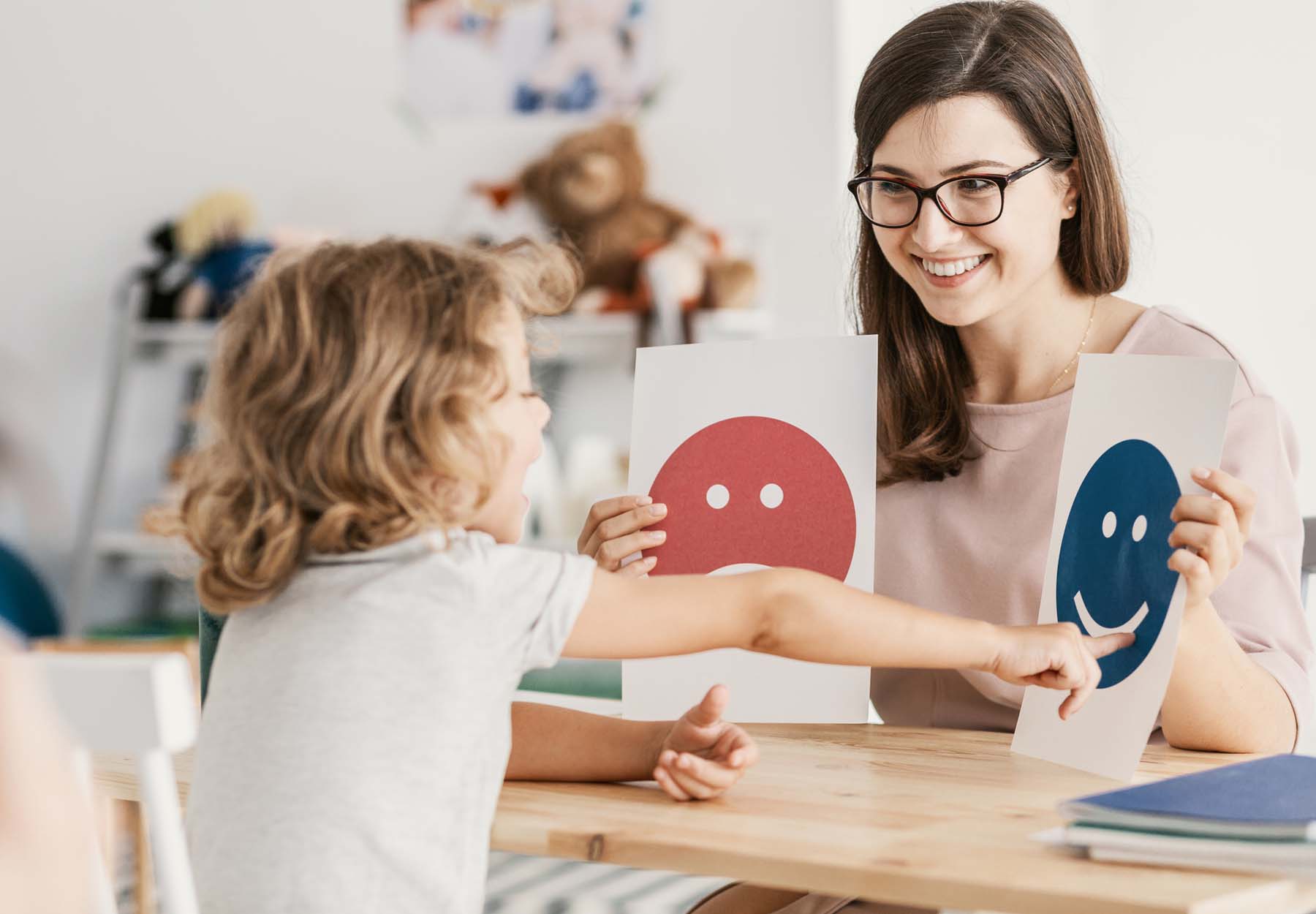 An autistic child points to an icon showing a happy face that is being held by a teacher.