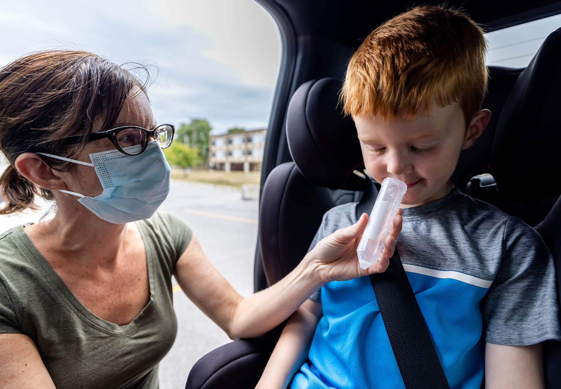 A mother, a nurse or a doctor is doing a Covid-19 saliva test on a cute redhead boy in his car. The women is wearing a surgical mask to protect herself from the spread of this infectious disease. The child is spiting in a plastic container, diagnostic medical tool. The saliva will then be analyzed in a laboratory for the coronavirus.