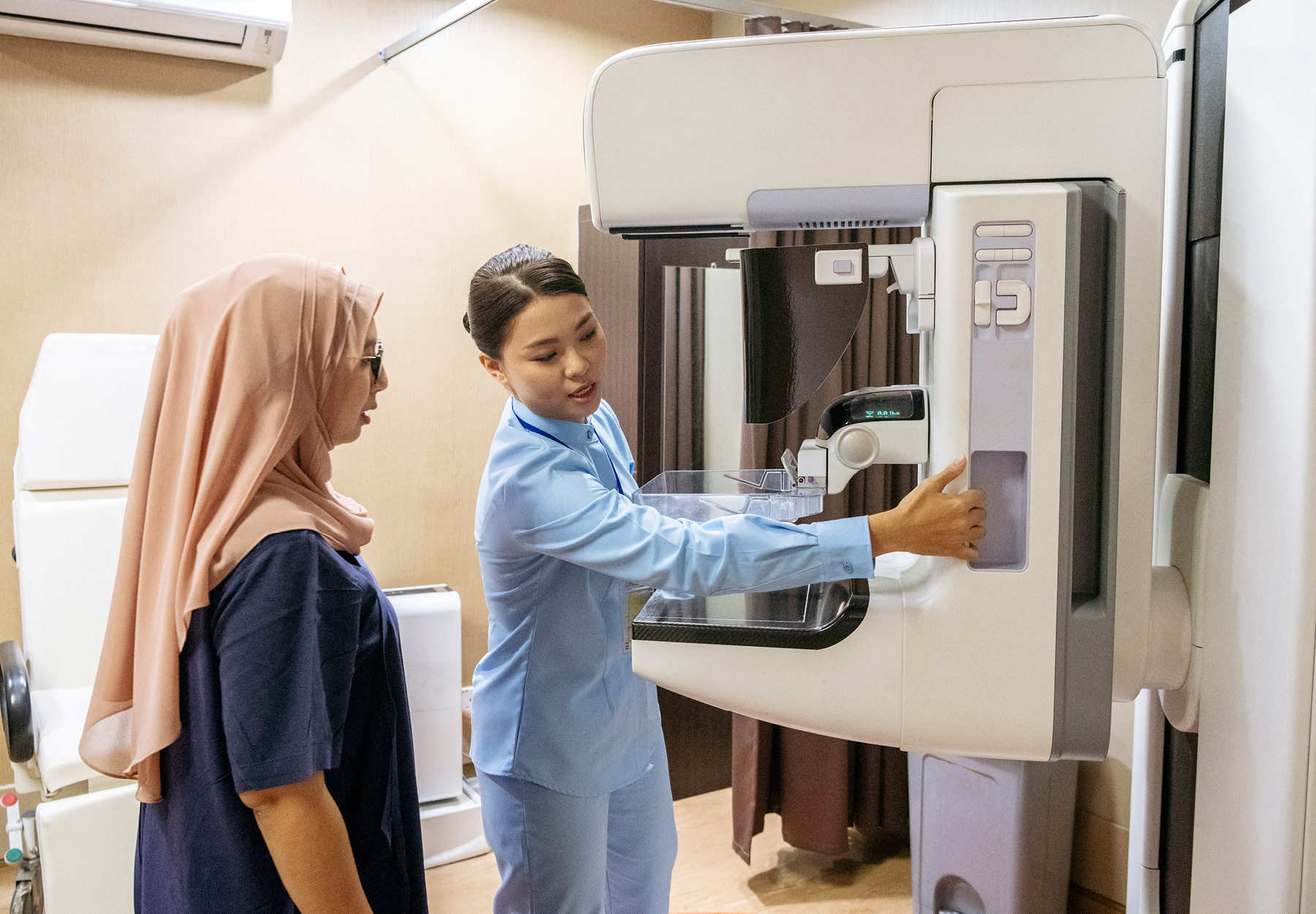 A technologist sets up a mammogram machine while a patient in a hijab looks on.