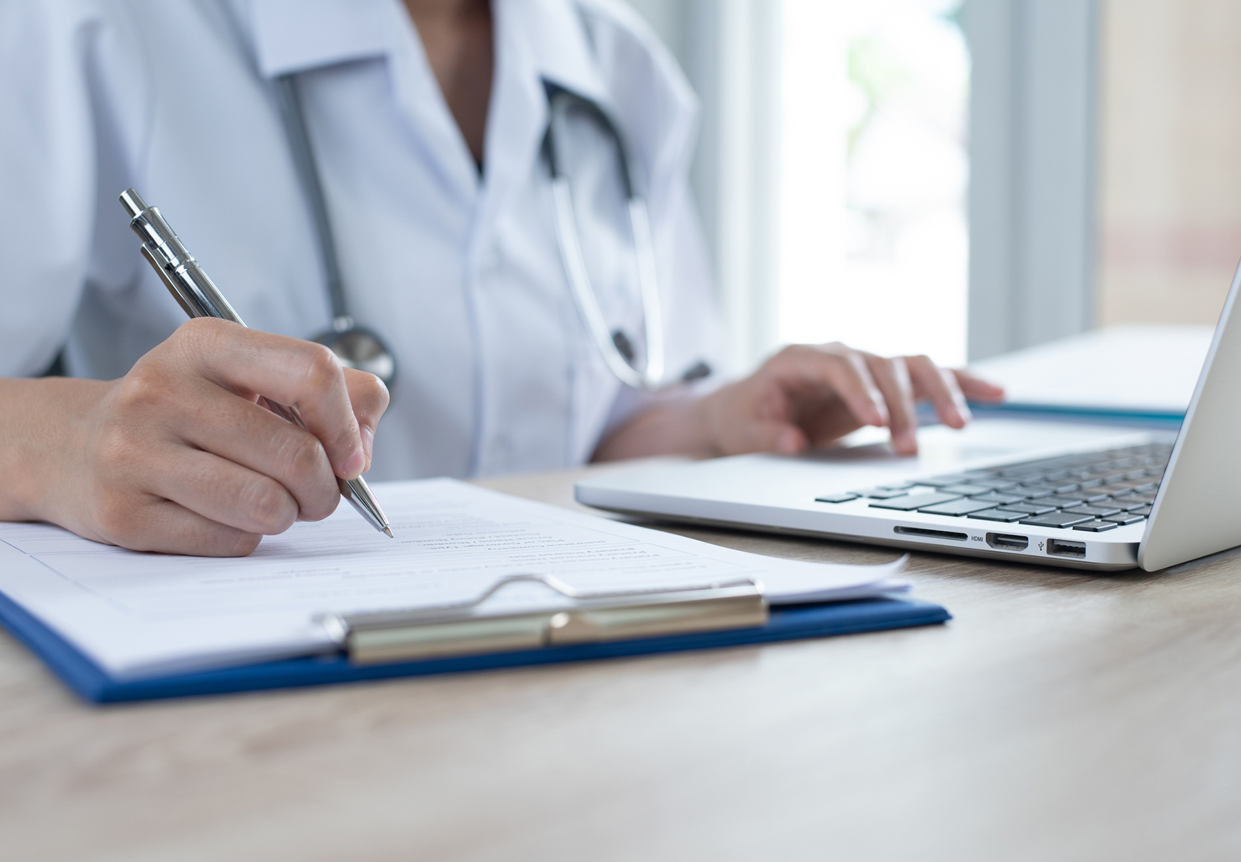 A closeup of a physician working on a laptop while writing on a clipboard.