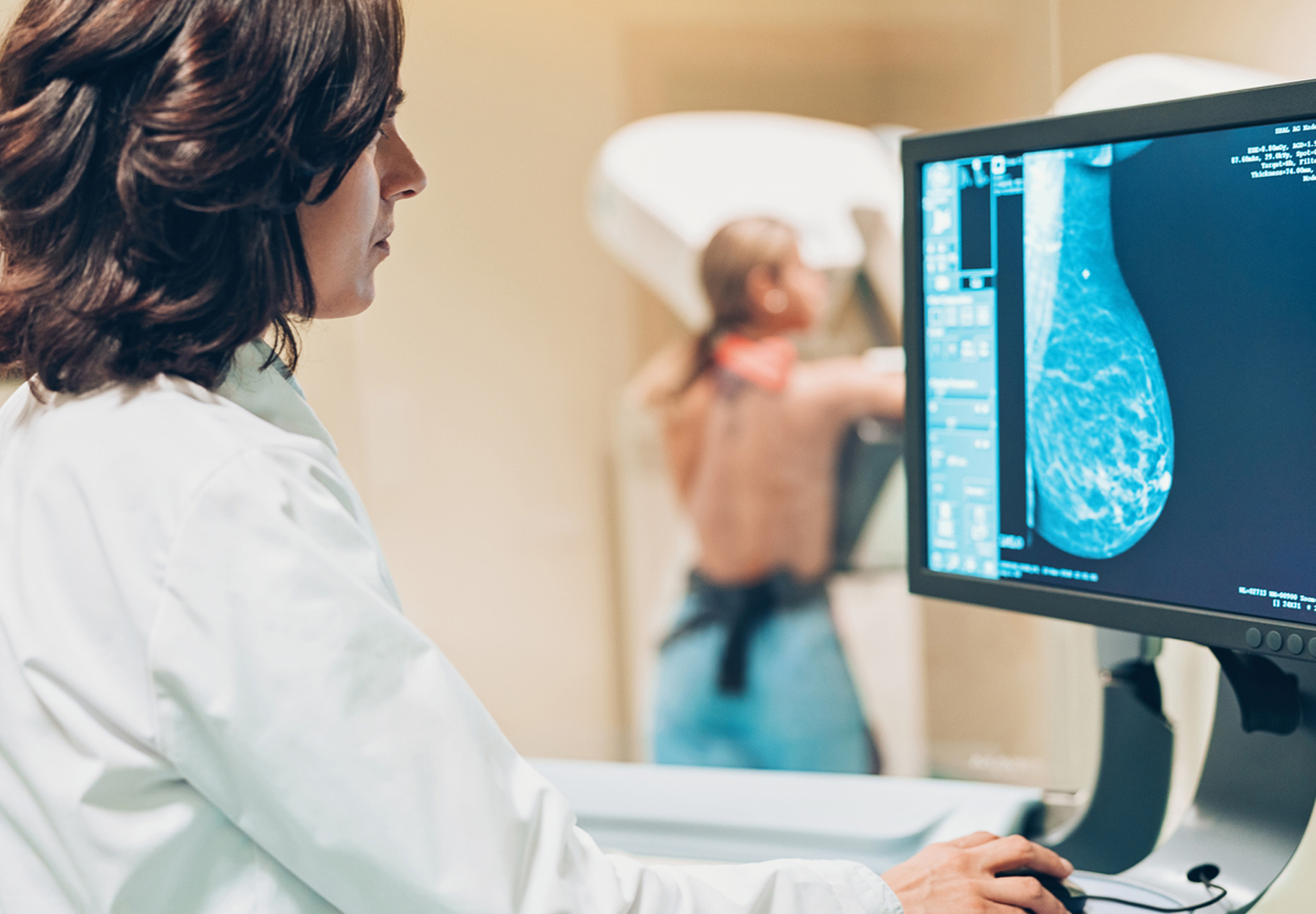 A medical technician looks at a computer screen while a patient stands in a mammogram machine in the background