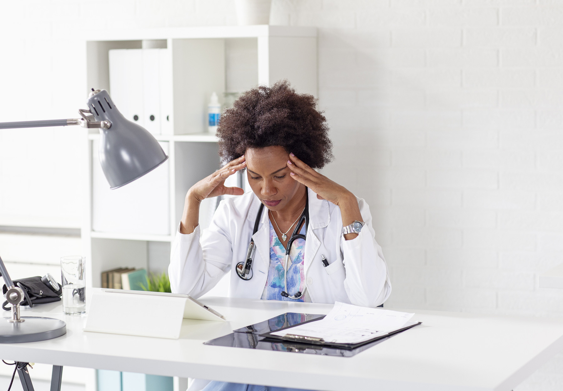 A stressed Black doctor sitting at her desk with her hands on her head.