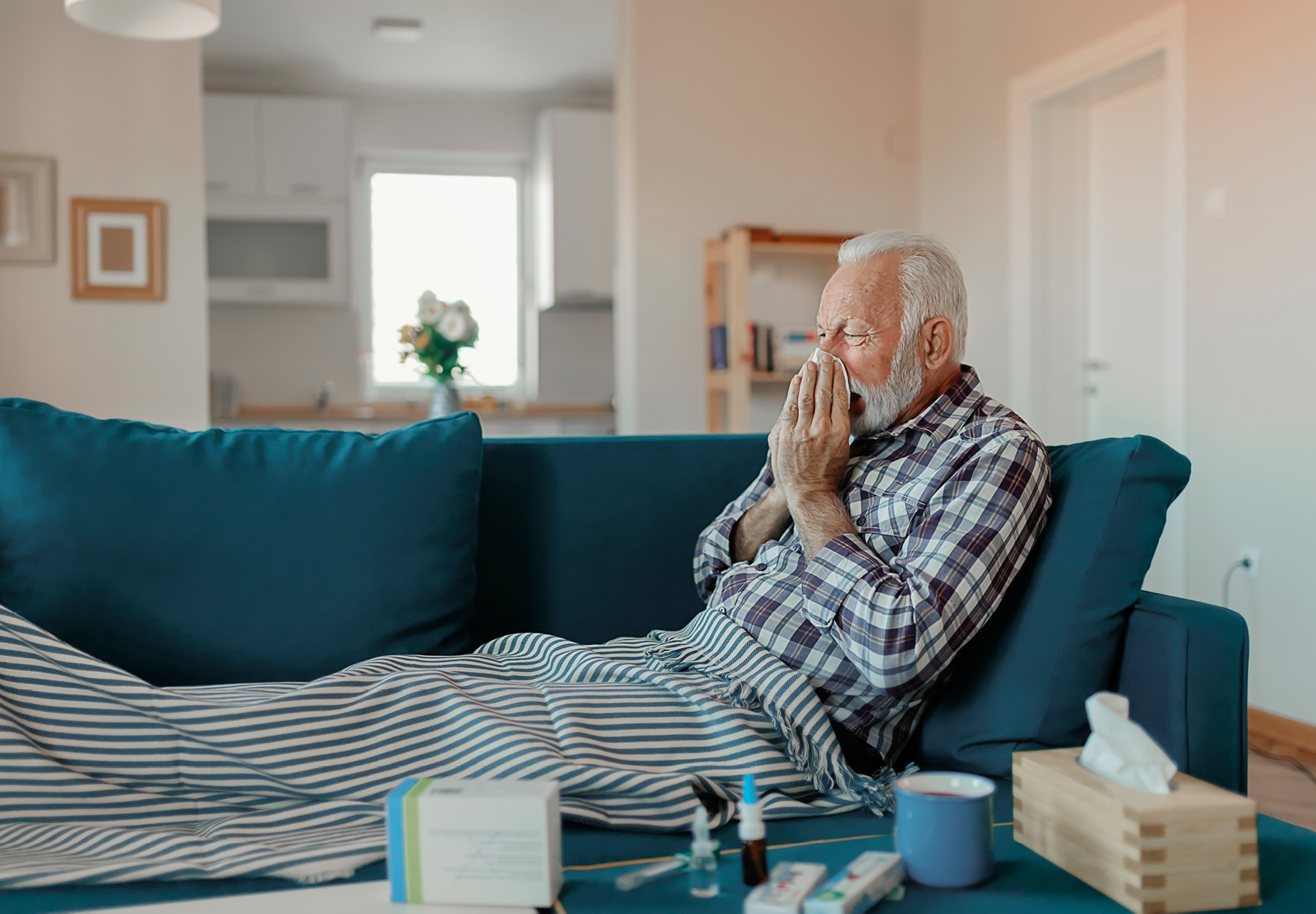 An elderly man sitting on the couch and blowing his nose.