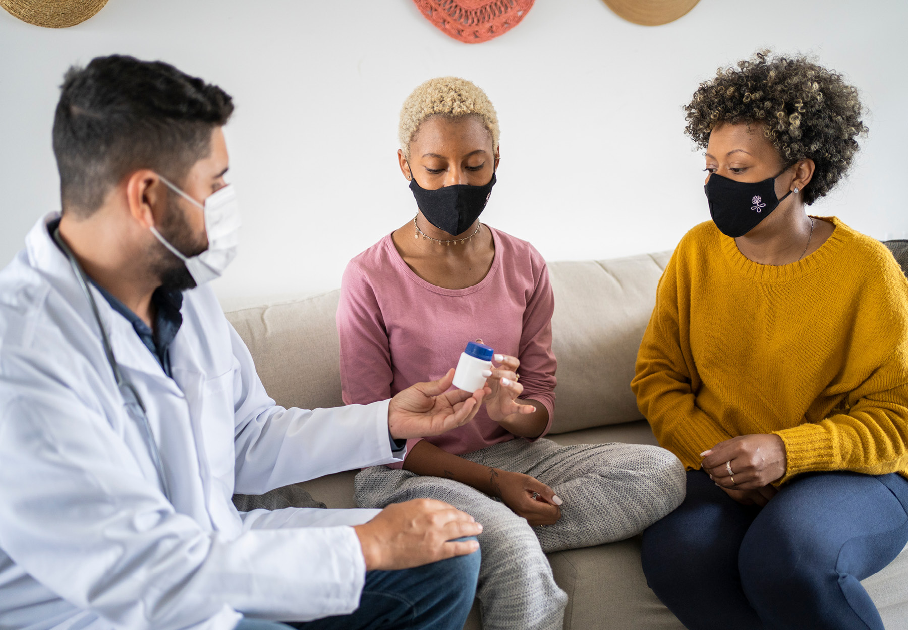 A young male doctor sitting with two young female patients hands one patient a bottle of pills. All are masked due to the COVID-19 pandemic.