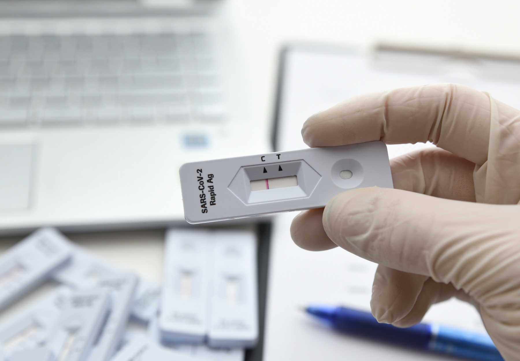 A closeup of someone holding a COVID-19 rapid antigen test cartridge in their white-gloved hand with a desk and laptop in the background to show a workplace setting.