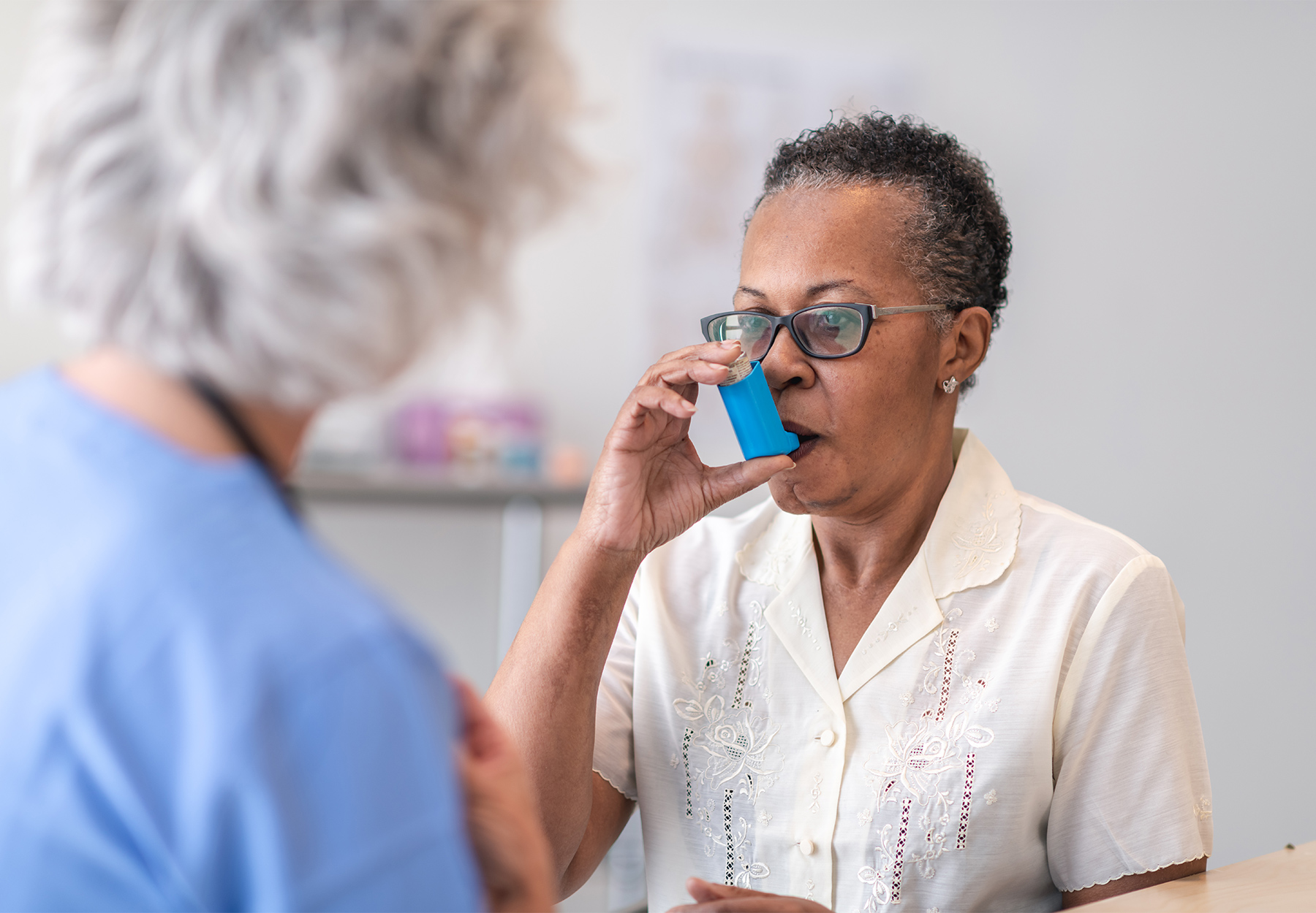 An elderly black woman uses an inhaler as her health care provider looks on.