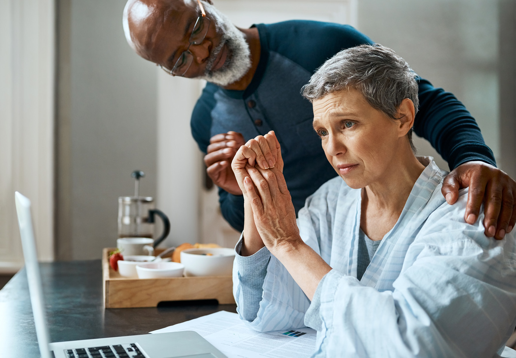 An elderly African American man comforting an elderly white woman who is sitting in front of a laptop