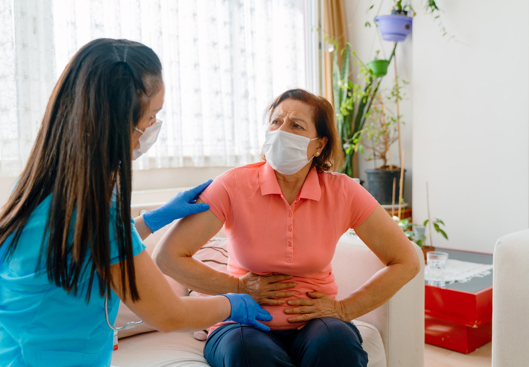 An elderly female patient wearing a mask holds her stomach in pain while a masked healthcare worker helps her, stock image