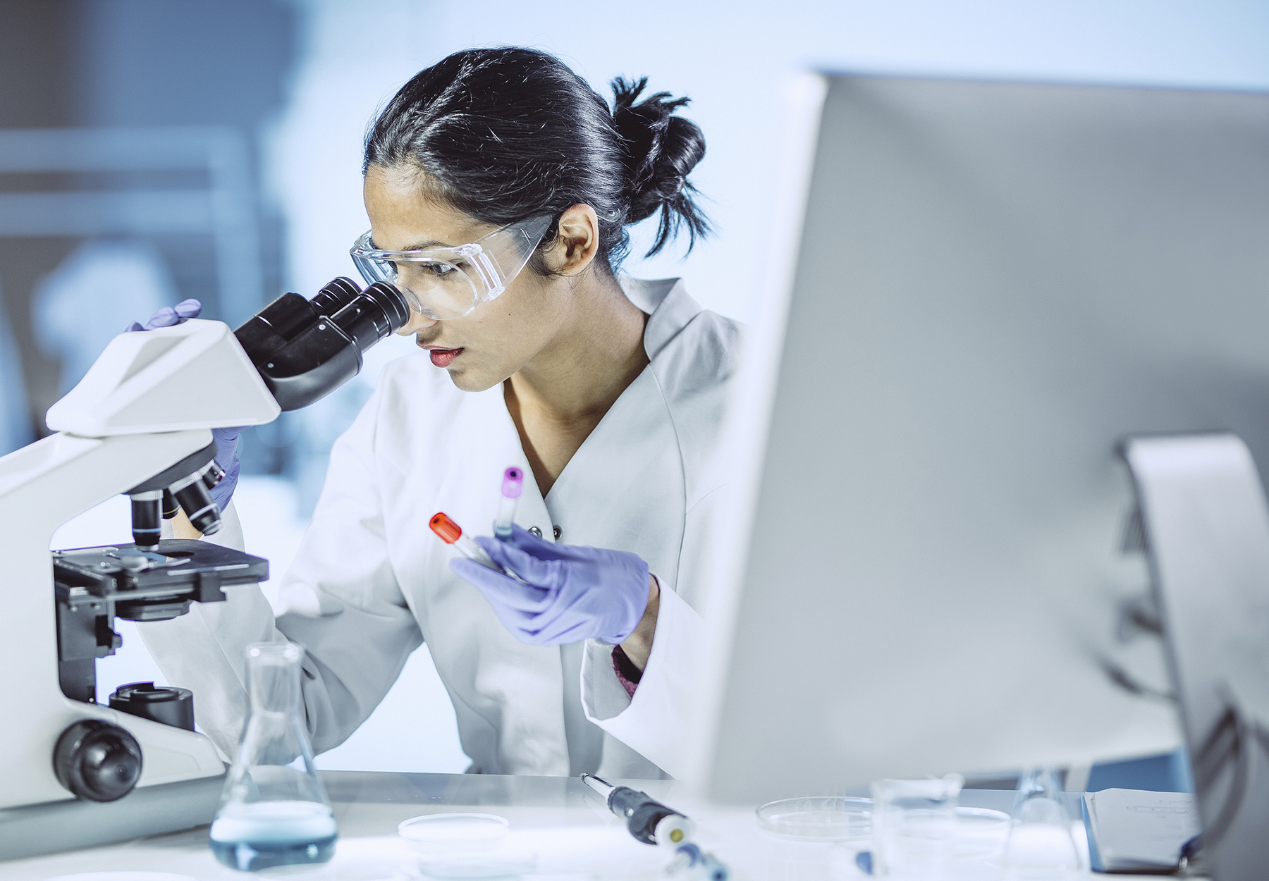 A female scientist working in the laboratory at a microscope while holding test tubes with samples. Stock image.