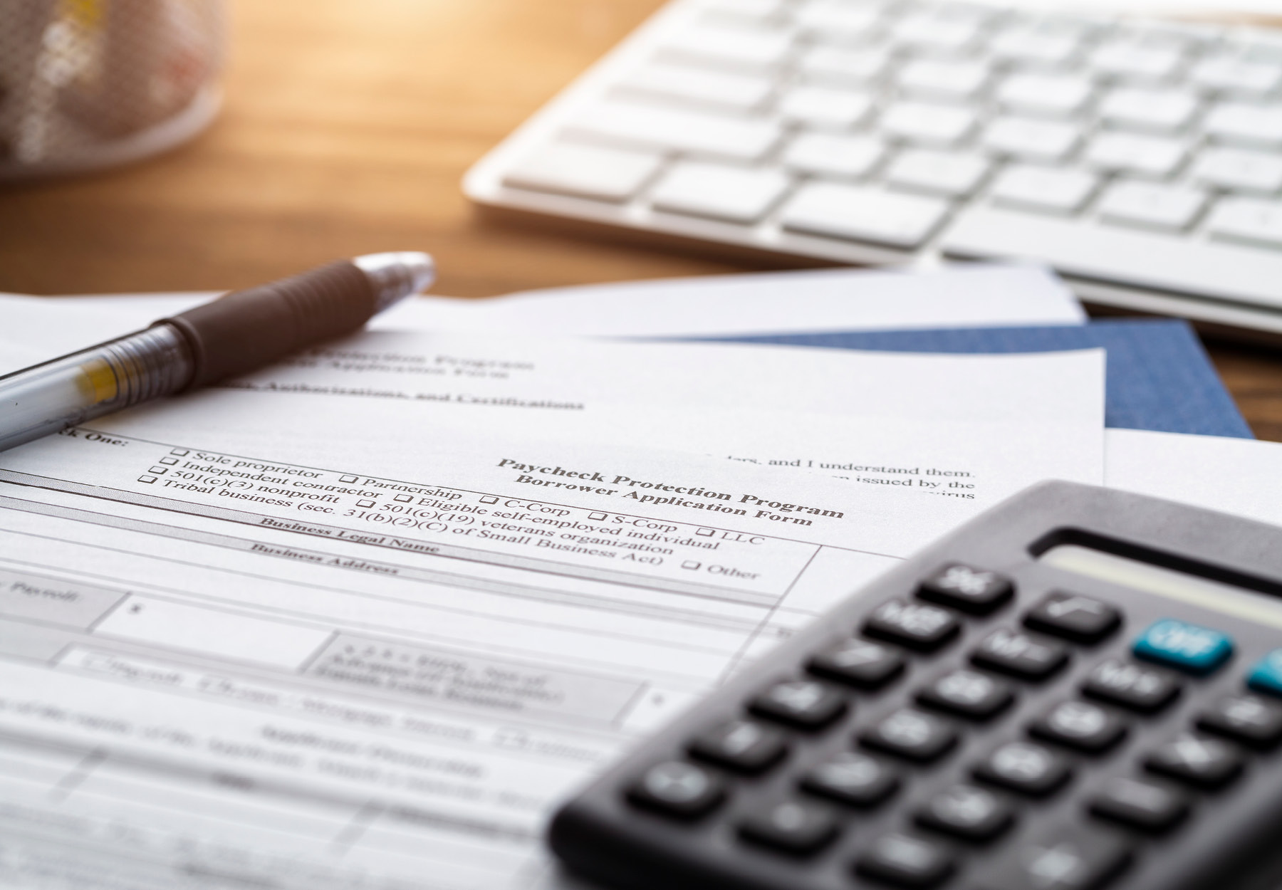 A closeup image of a desk with a Paycheck Protection Plan document, computer keyboard, pen, and calculator on it.