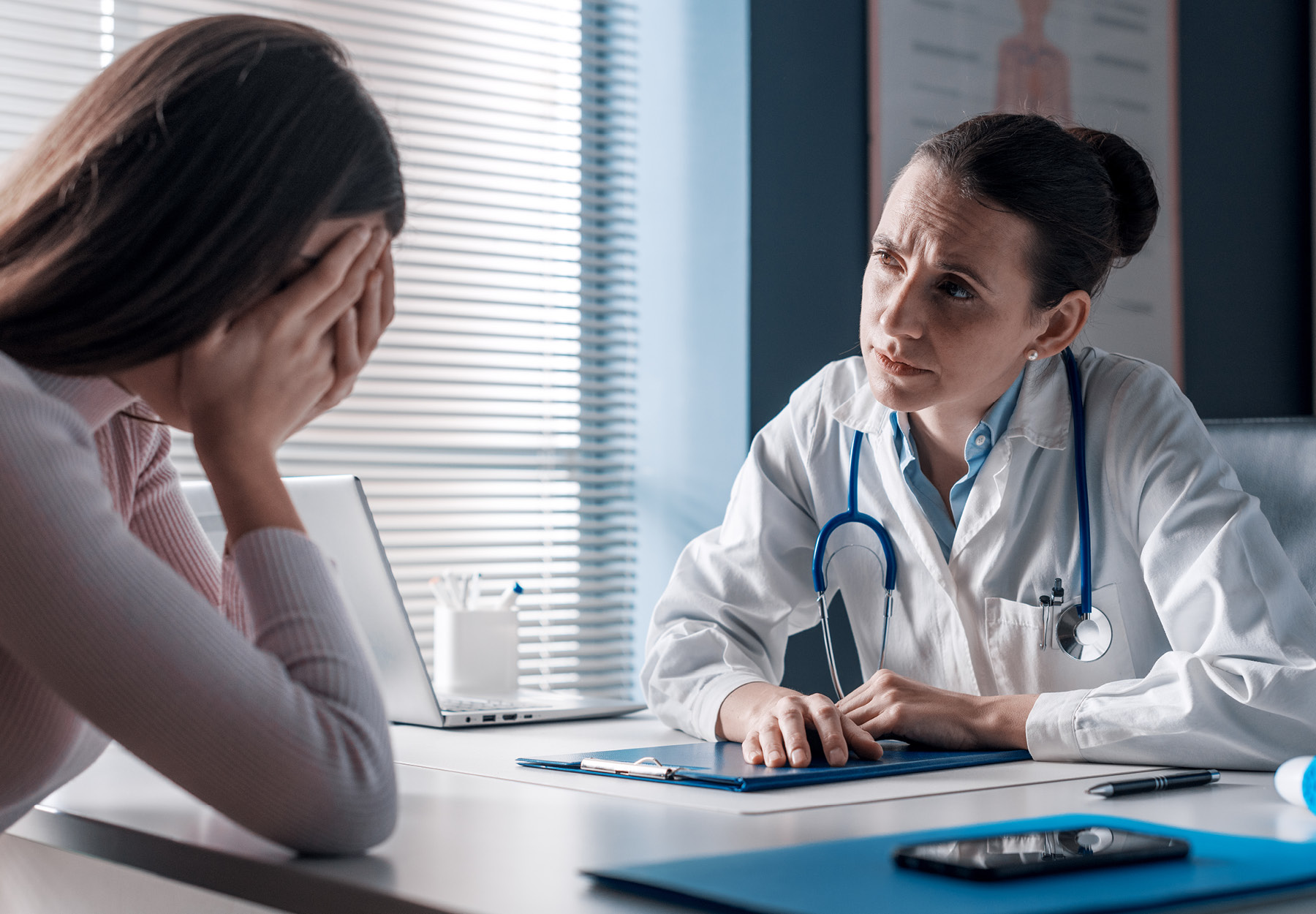 A doctor frowns in concern at an upset patient who has her head in her hands.