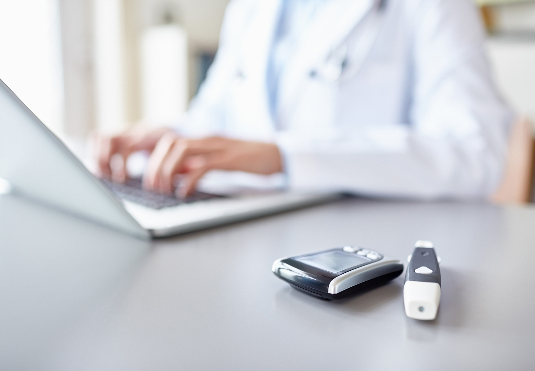 Close-up of glucometers on desk with female doctor using laptop in background wearing lab coat and stethoscope. Stock image.