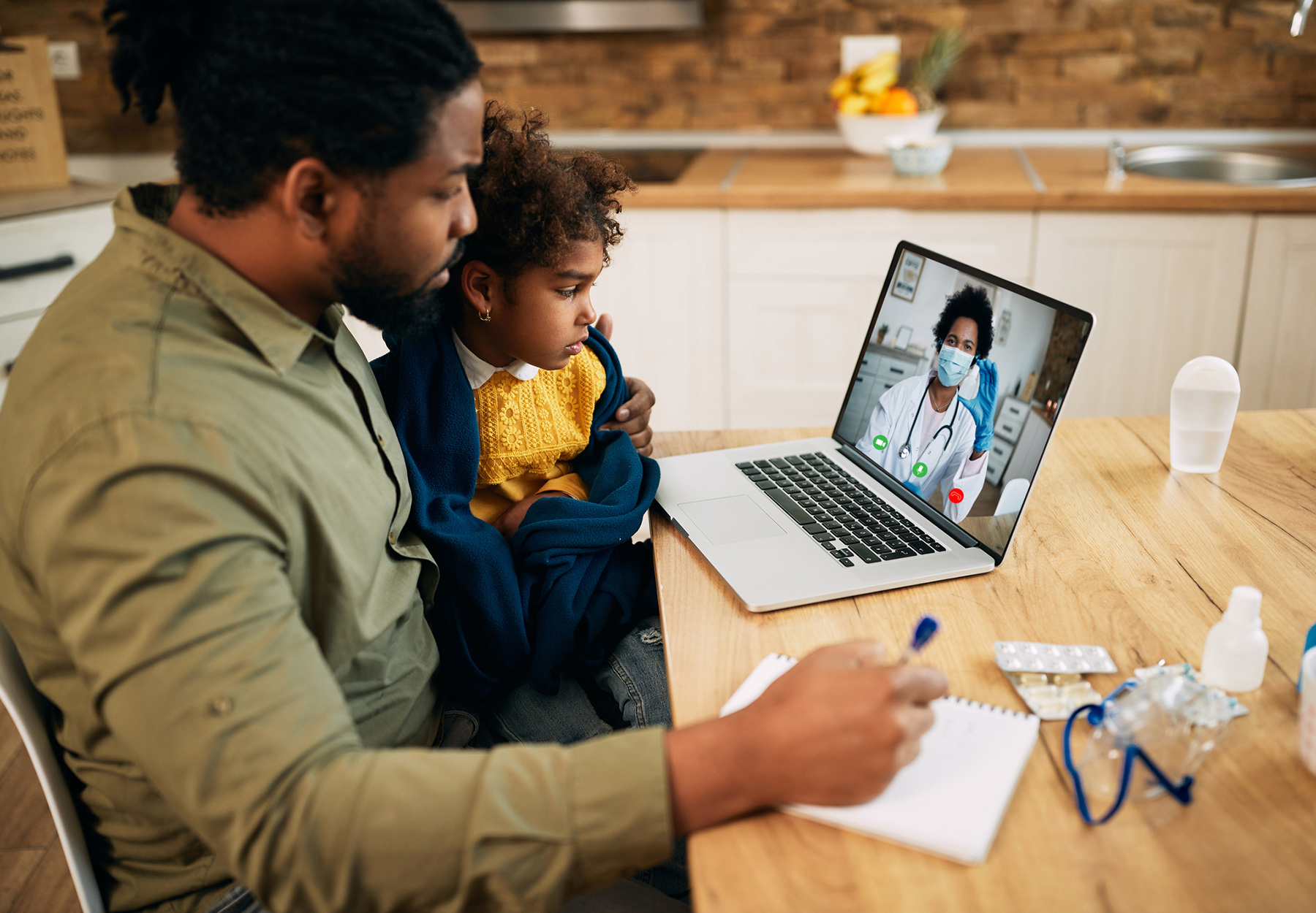 Stock image of Black father and daughter communicating with a doctor via video call from home during COVID-19 pandemic. Focus is on girl.