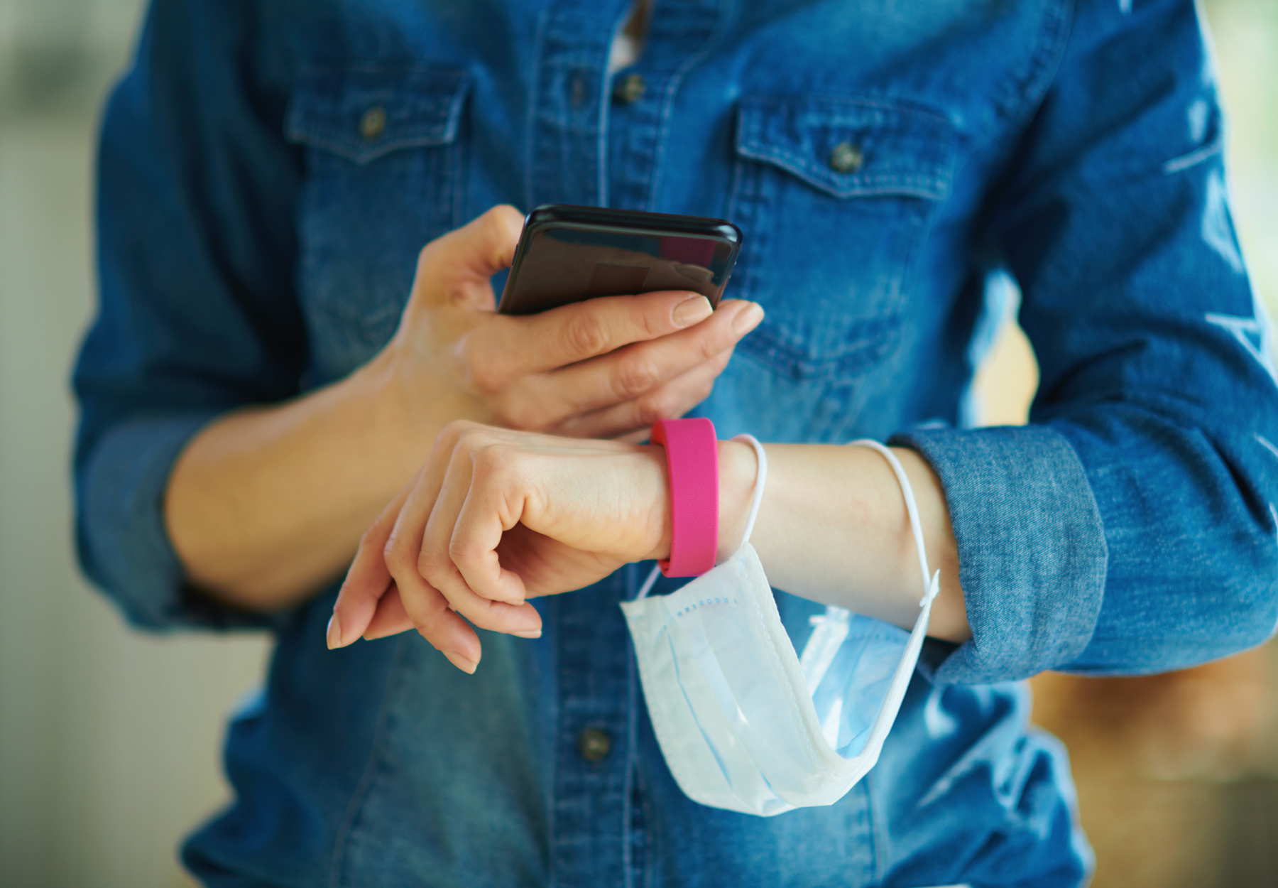 Closeup stock photo of woman wearing a bracelet-type smart device while checking her smartphone and holding a medical mask