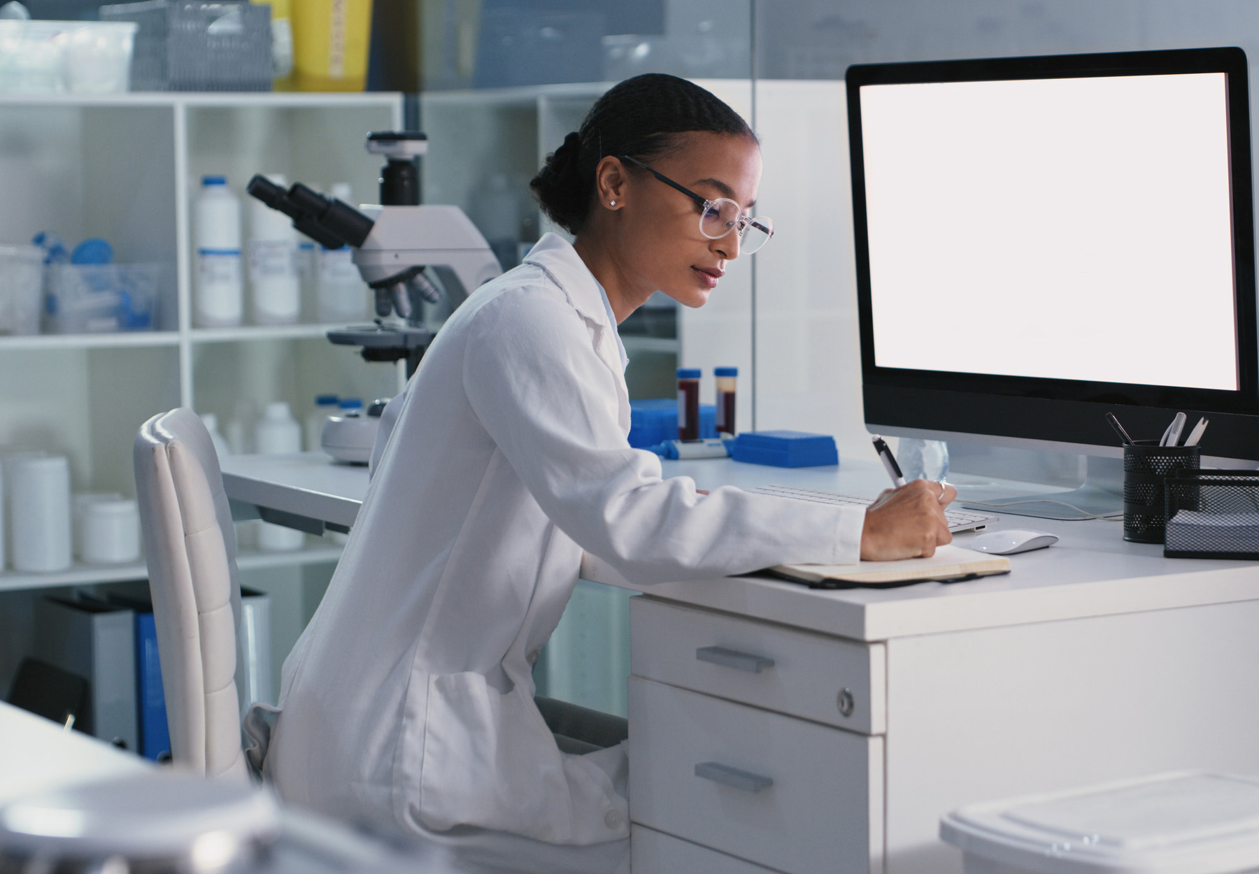 A female lab worker sits at her desk, writing in a notebook. There is a computer screen in front of her. Stock image,