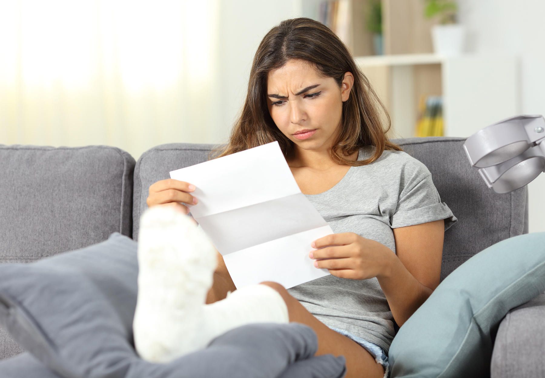 Woman with a cast on her leg is sitting on a couch looking at a medical bill and frowning. Stock photo.