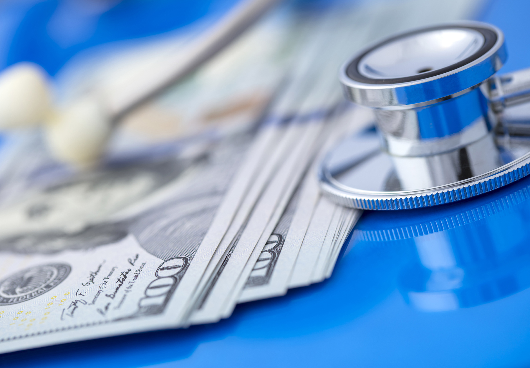 Close-up of a stethoscope with US $100 dollars bills on a blue desk. Shallow depth of field.