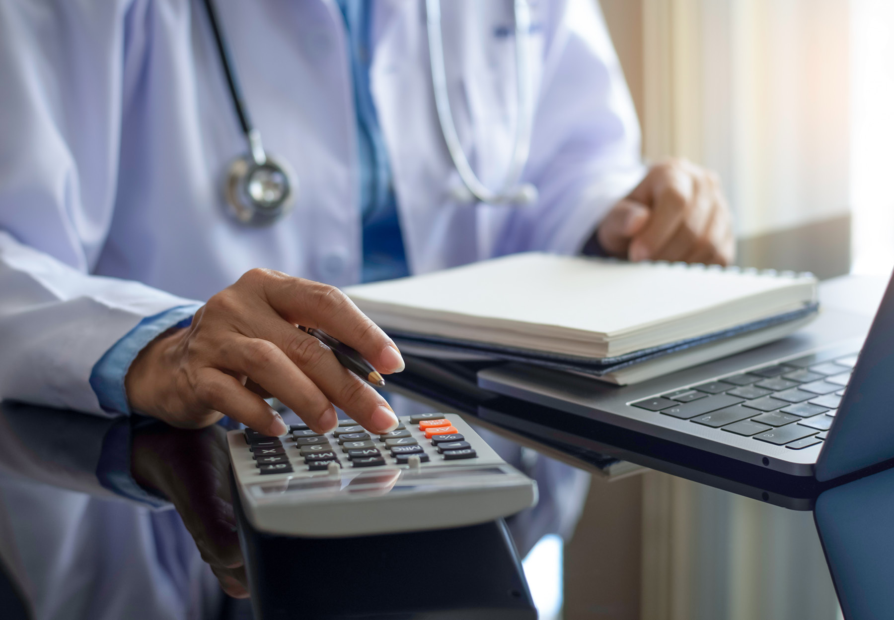 Female doctor or physician with stethoscope using calculator and work on computer notebook on the desk at modern office at clinic or hospital. Stock photo.