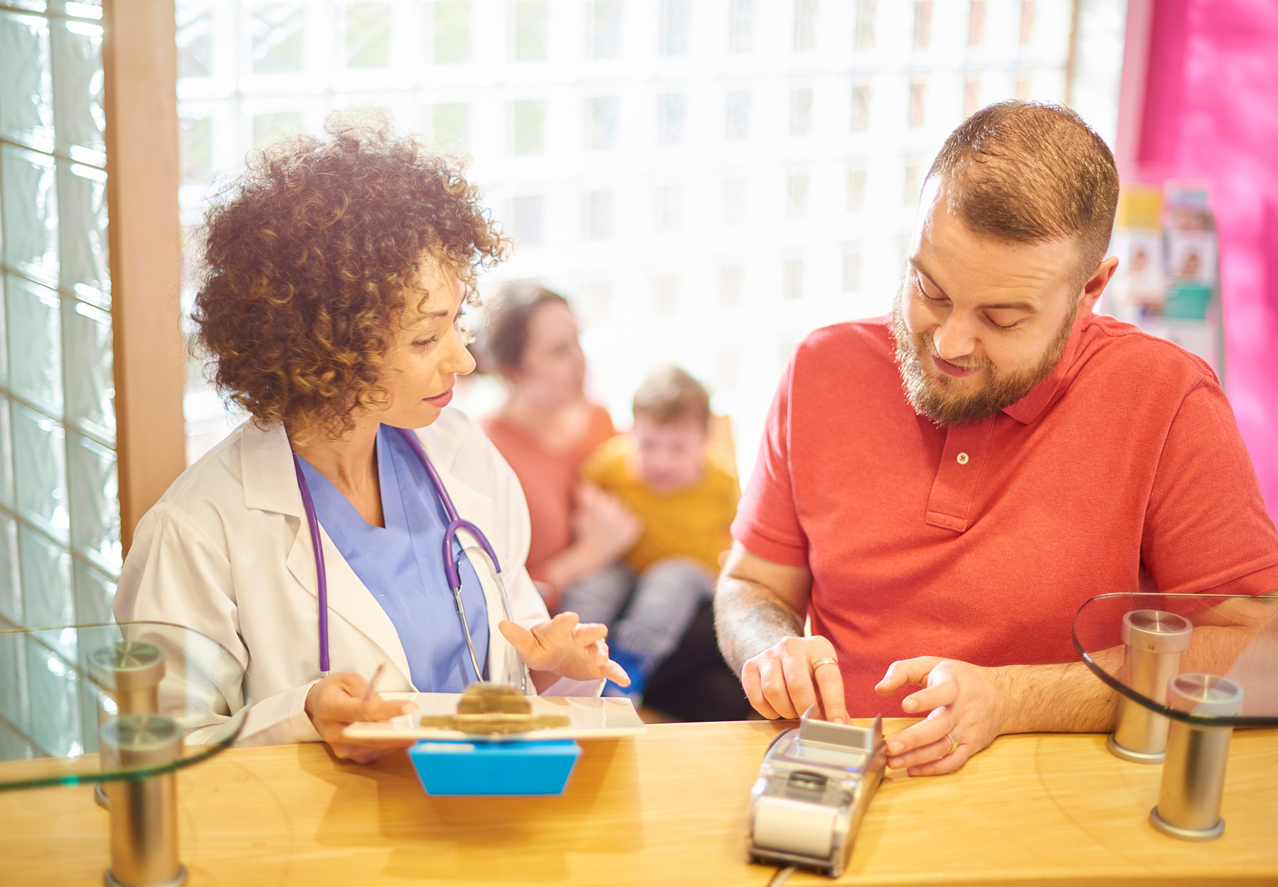 A male patient pays his medical bill at the reception desk with his family sat in the background. Stock photo.