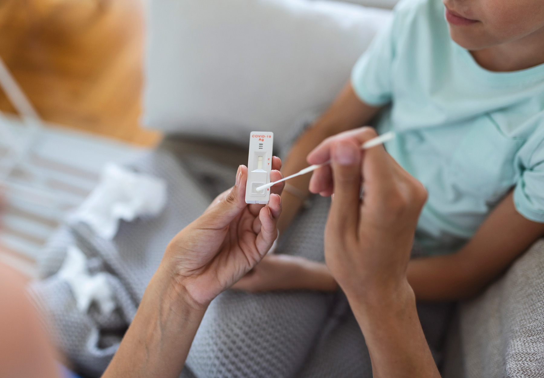 Closeup of mother performing at-home COVID-19 antigen test for her daughter. The mother is holding the swab and test cartridge while the daughter sits in the background. Stock photo.