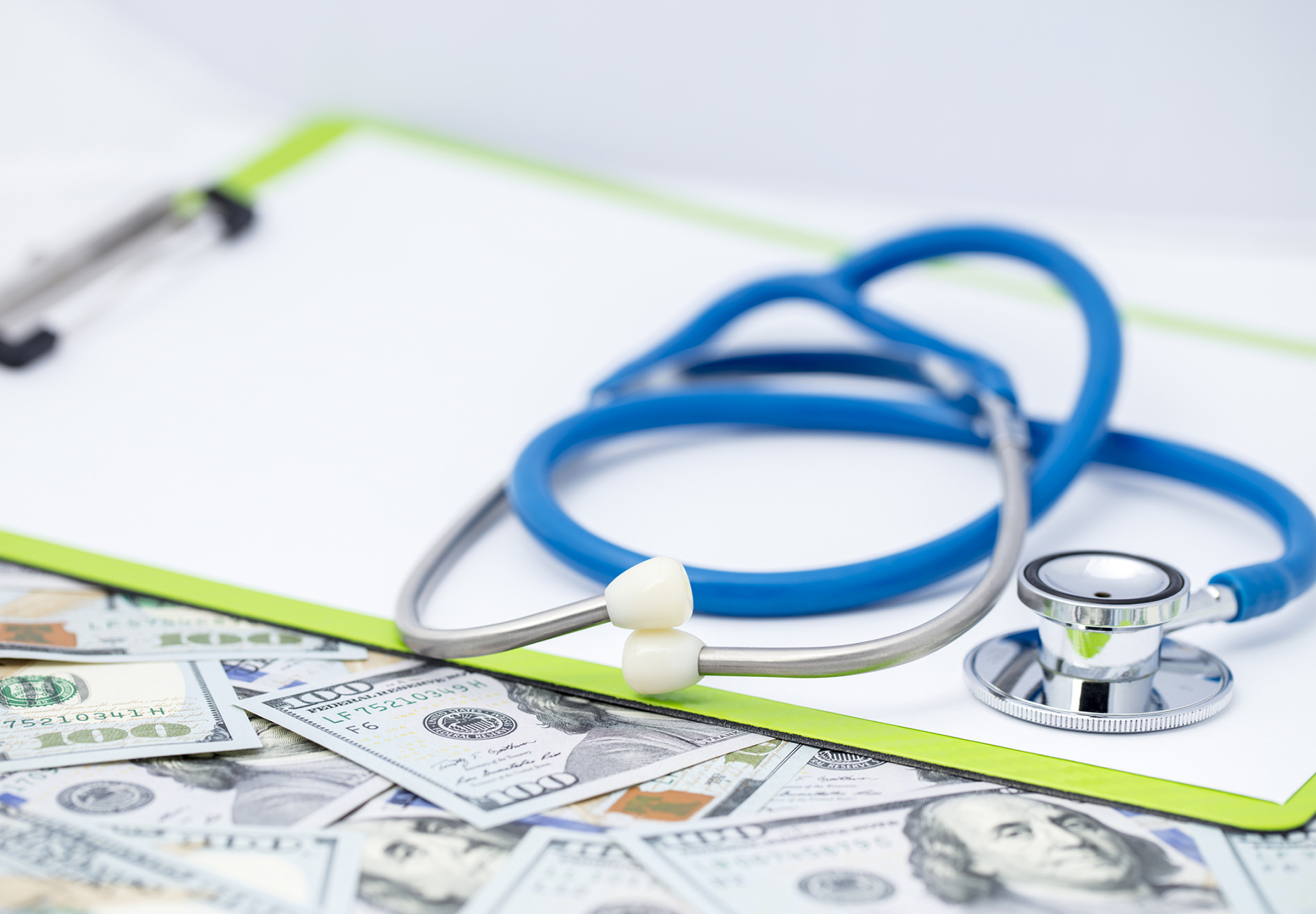 Close-up of a stethoscope and a clipboard on a heap US $100 dollars bills. Shallow depth of field. Stock photo.