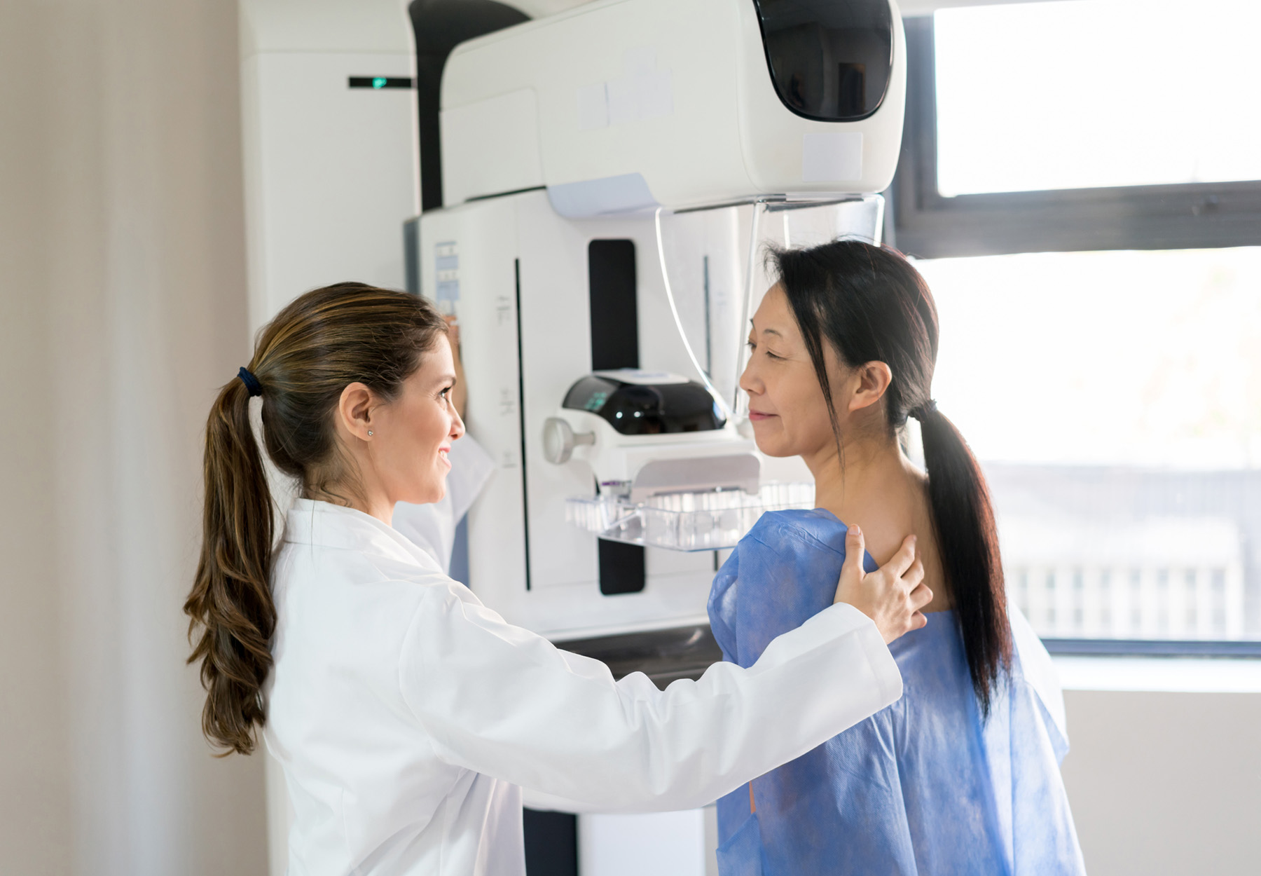 Friendly female doctor talking to her patient and adjusting her position to do a mammogram at the clinic. Stock photo.