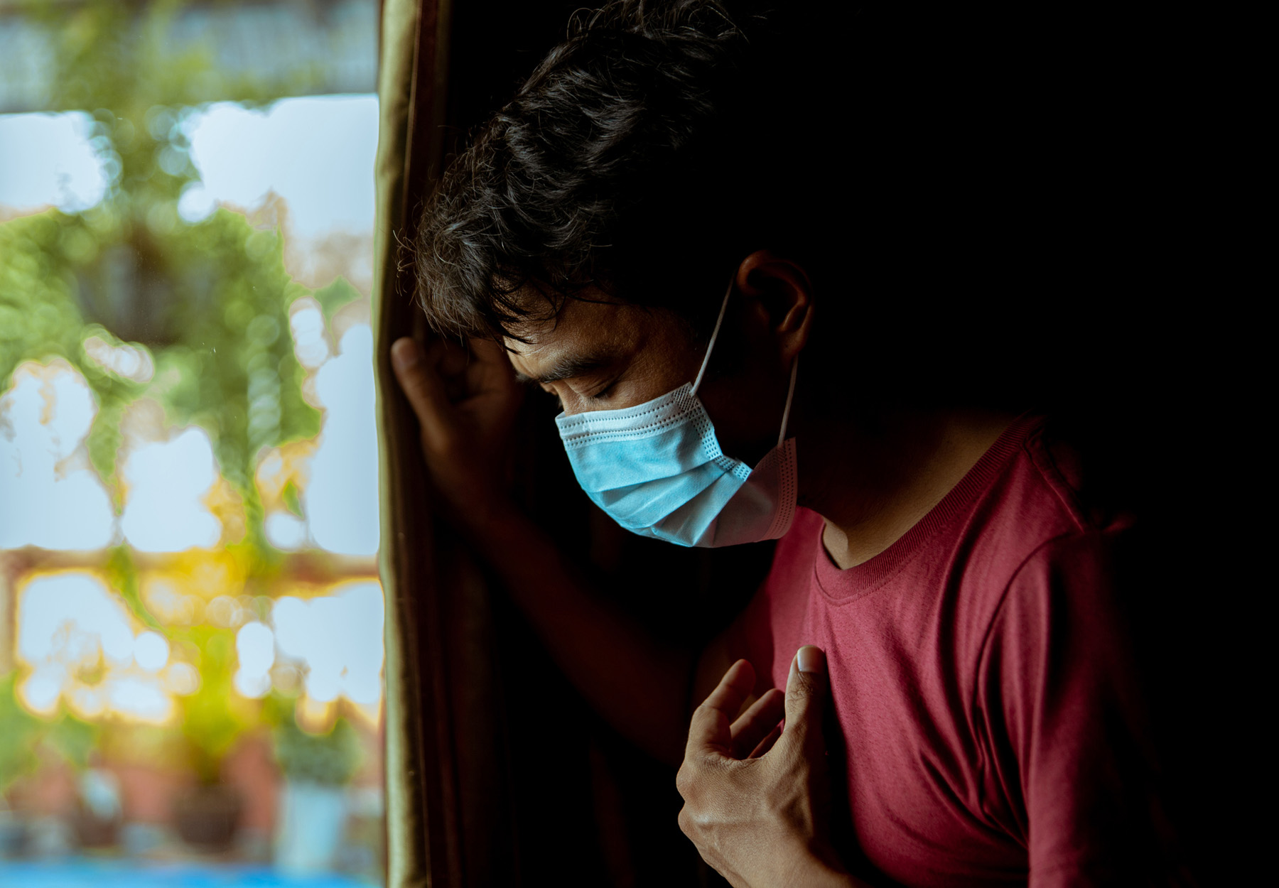Man with medical mask slumped at a window while gripping his chest. Long COVID concept. Stock photo.