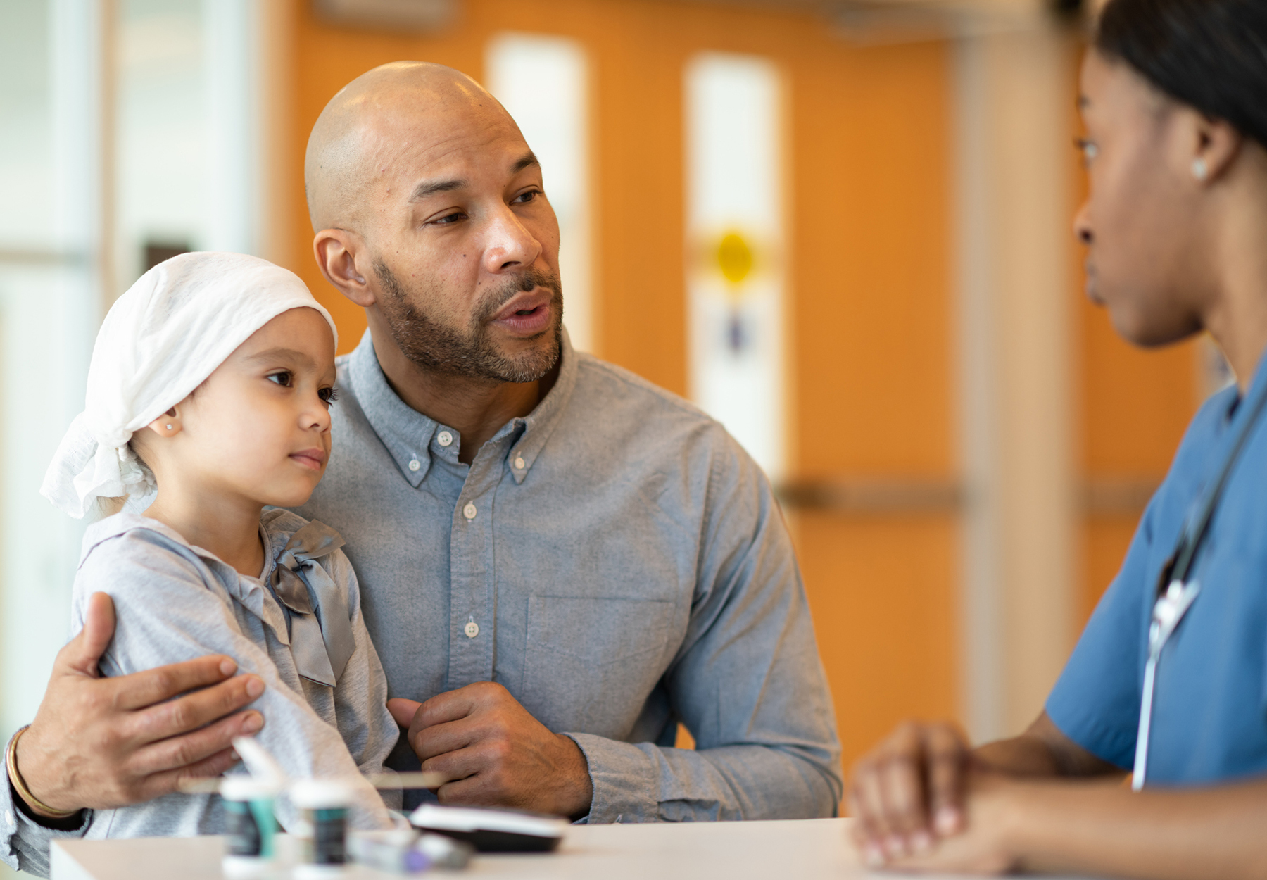 An African American father accompanies his young daughter to a medical appointment. His daughter has cancer and she is wearing s headscarf. The two are seated at a table across from a medical professional. The black female doctor is sharing information about the girl's course of treatment. Stock photo.