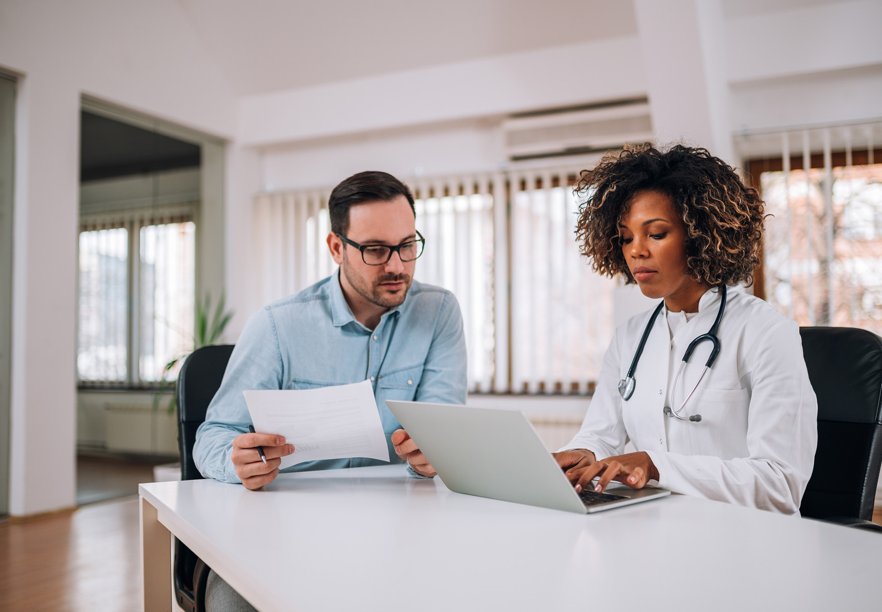 Doctor meeting with medical employee at a desk. The doctor is typing on her laptop and the employee is holding a piece of paper. Stock photo.