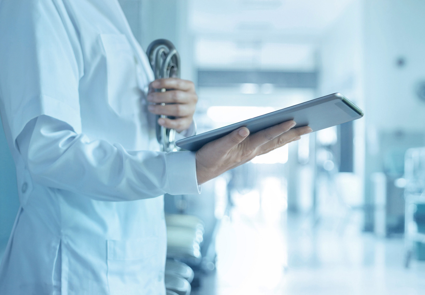 A closeup of a healthcare professional in a white coat holding a tablet and stethoscope. The image has blue tones. Stock photo.