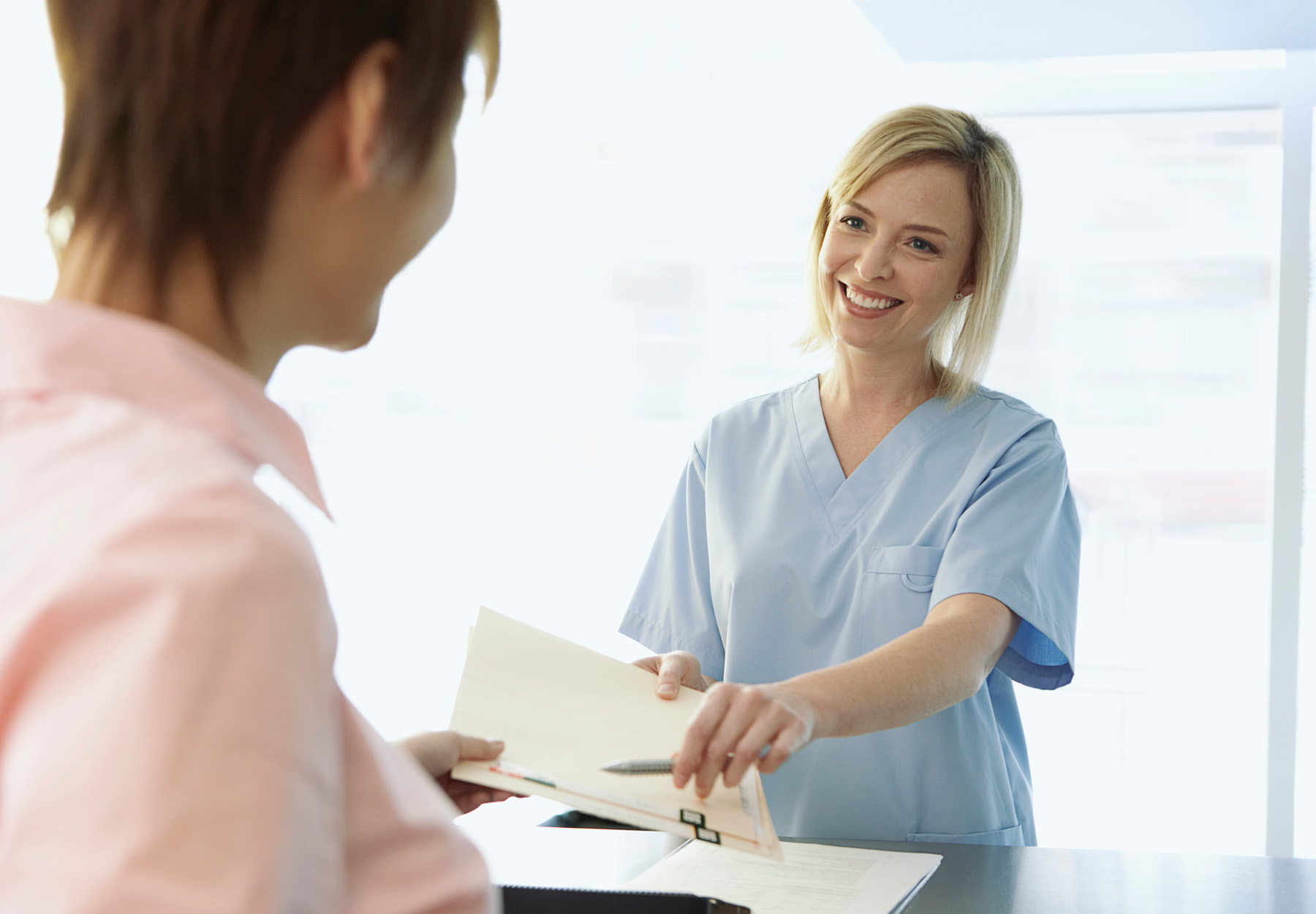 A female healthcare office assistant in light blue scrubs hands medical records and a pen to a female patient. Stock photo.