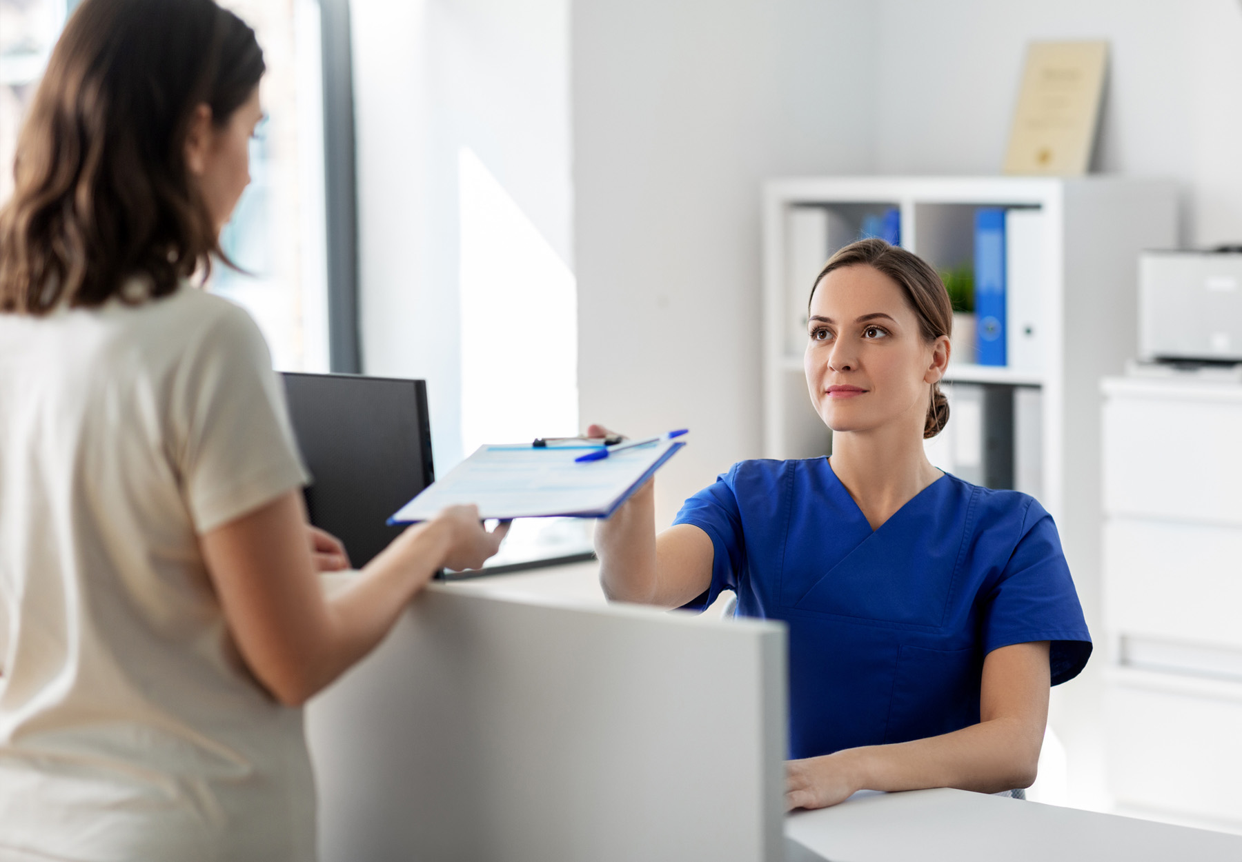 Healthcare professional in blue scrubs sits at her desk and hands a clipboard with a form on it to a female patient. Stock photo.