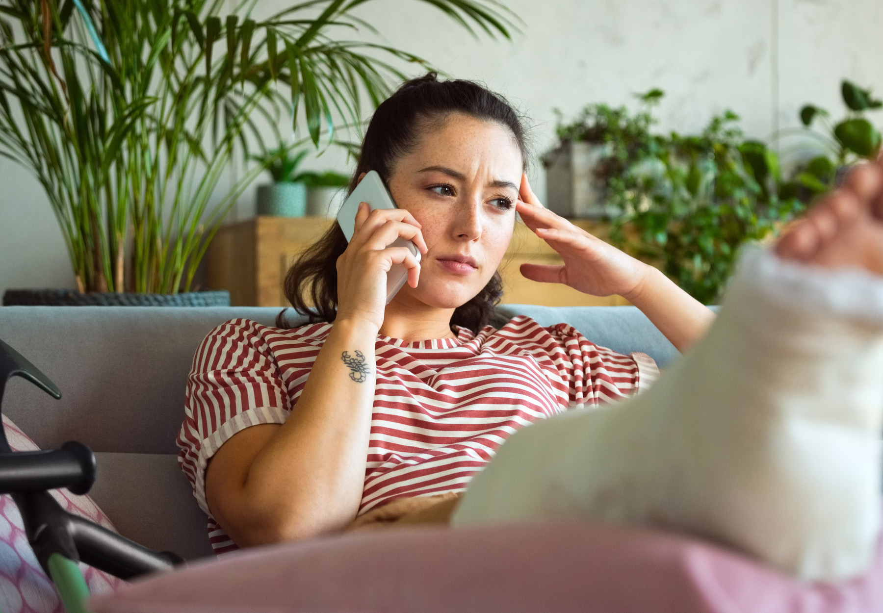 A woman sitting on the couch with her leg propped up in a cast. She is on her cellphone and has a worried look on her face as she just got a surprise medical bill. Stock photo.