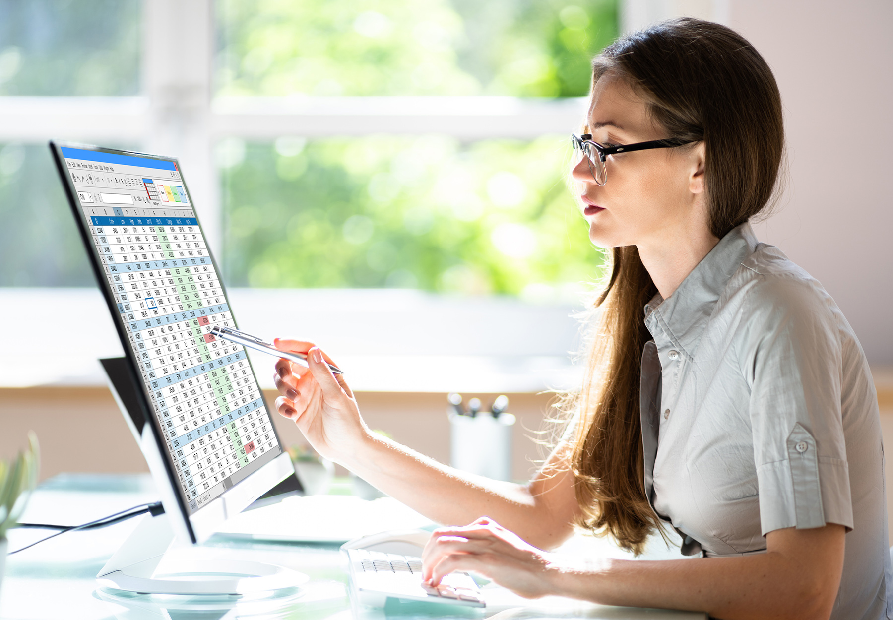 Female lab employee with glasses looks at spreadsheet on a computer monitoring. She is pointing something out with a pencil on the screen. Stock photo.