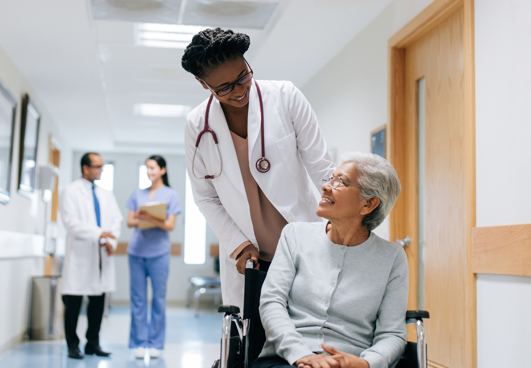 African American doctor pushes an elderly patient of Asian descent in a wheelchair down a hospital hallway. Two hospital staff members can be seen in the background. Stock photo.