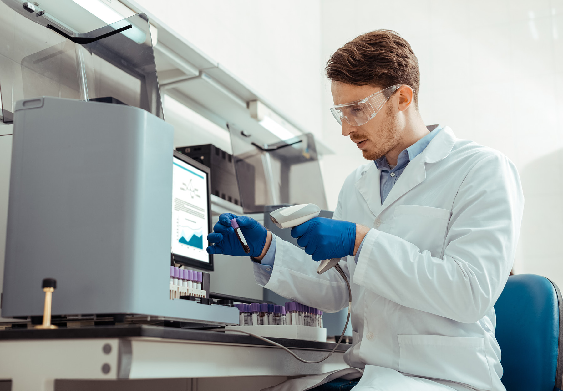 White male lab employee in PPE scans a test tube of blood while running tests in the lab.