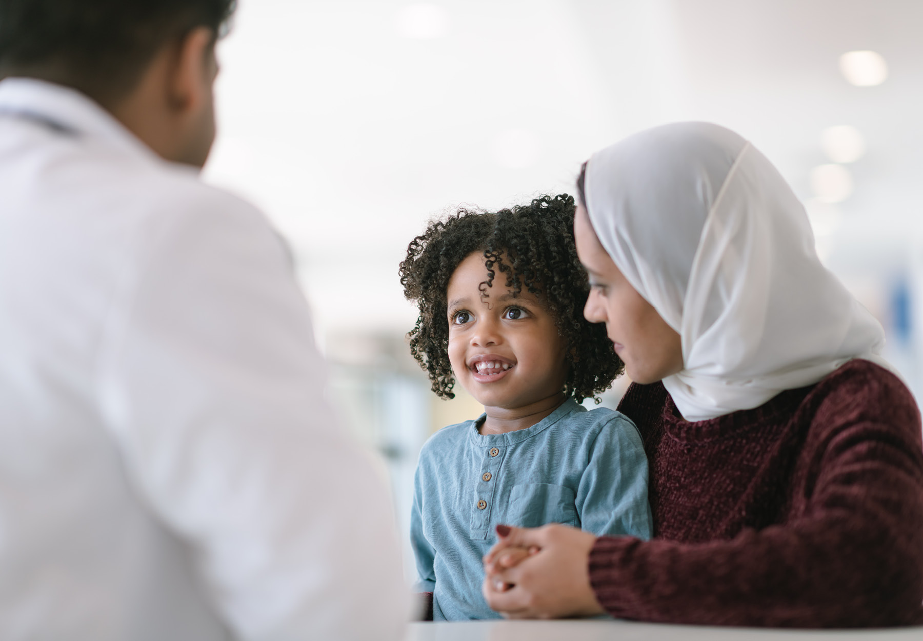 A young Muslim mother wearing a hijab takes her toddler son to visit the pediatrician. The boy is sitting on his mother's lap. They are seated at a table and a male doctor is seated across from them. The child is smiling at the doctor. The mother is looking at her son. Stock photo.