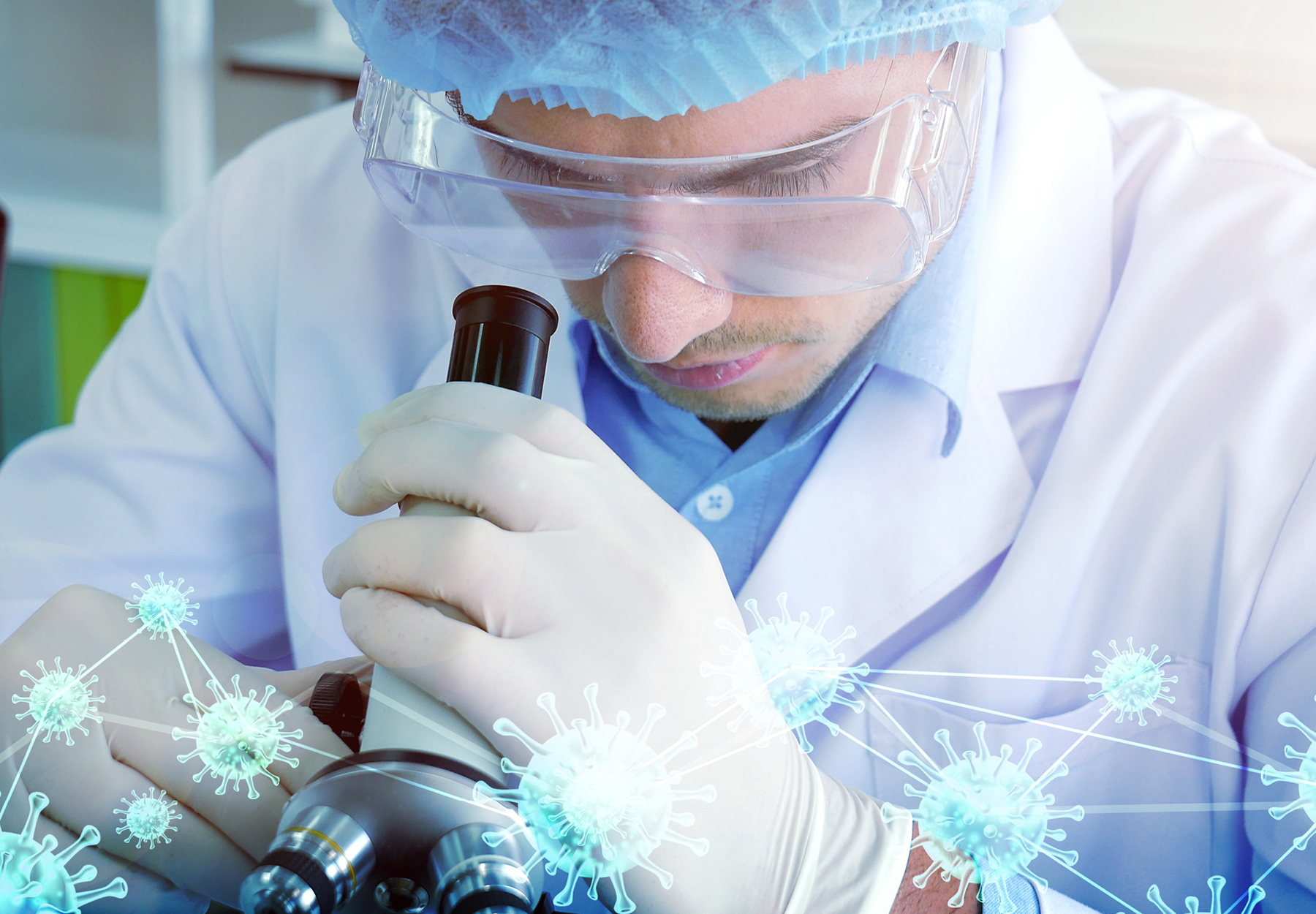 An image of a researcher in full PPE looking through a microscope with COVID-19 molecule graphics underneath to show the concept of COVID-19 test development. Stock image.