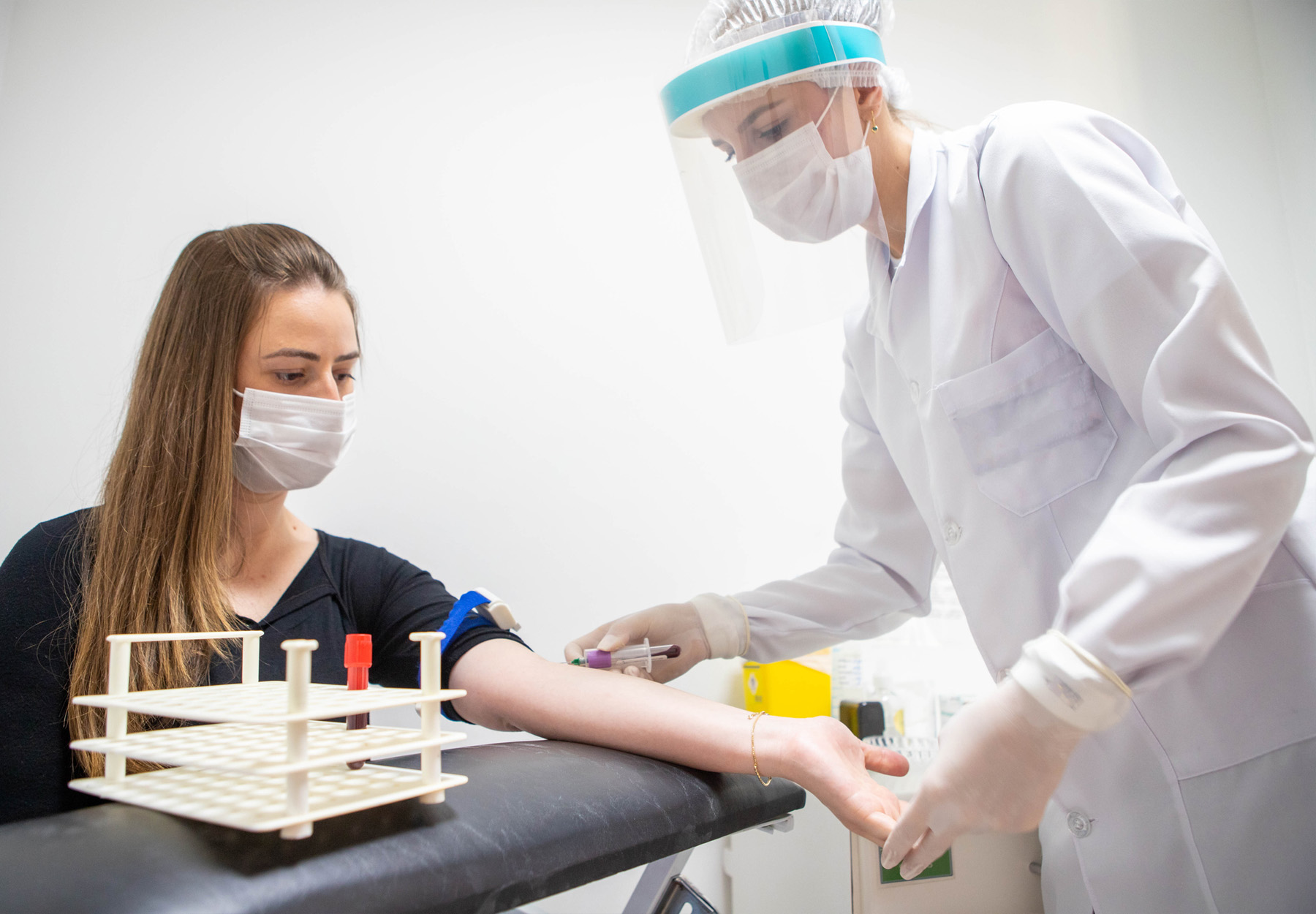 Young, white female healthcare worker in full PPE performing a blood test on a young, white female patient who is wearing a mask. Stock photo.