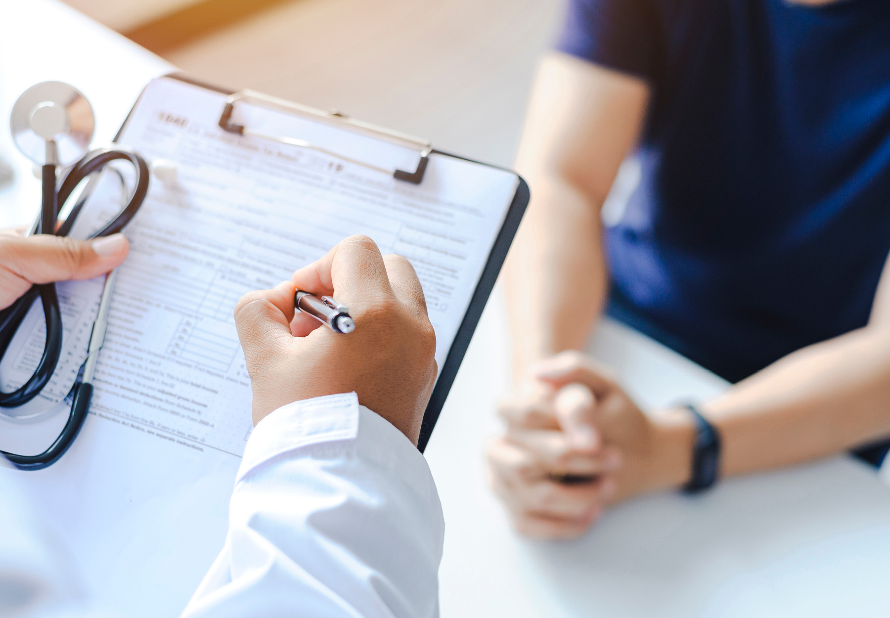 Closeup of doctor filling out a form on a clipboard at a desk with a healthcare working in scrubs sitting opposite. Stock photo.