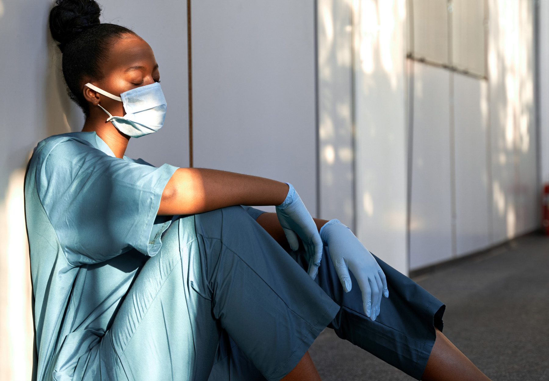 Stressed African American healthcare worker in scrubs, mask, and gloves, sits in the hallway of a hospital with her eyes closed. Stock photo.