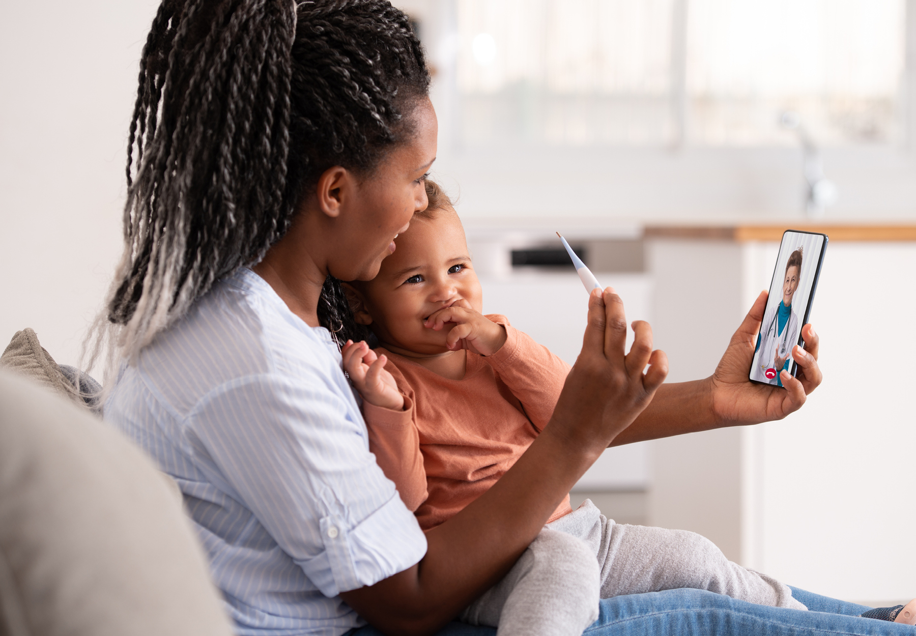 An African American mother with her toddler-age daughter on her lap, holds her phone in one hand and a thermometer in the other as she meets her doctor for a telemedicine appointment. Stock photo.