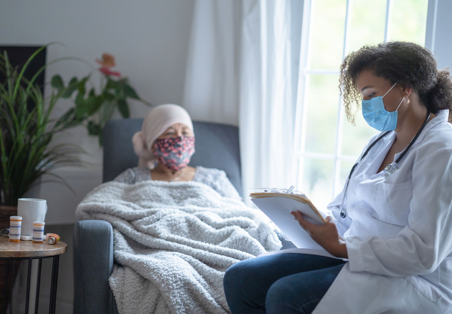 In a hospital room, an female cancer patient of Asian descent is chatting with her doctor. Both of them are wearing a mask during the coronavirus pandemic to help prevent the transfer of germs. The doctor is a female and of African American ethnicity.