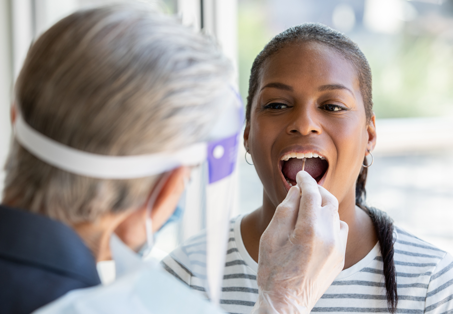 Medical worker with face shield taking a swab sample of a patient's throat. iStock image.