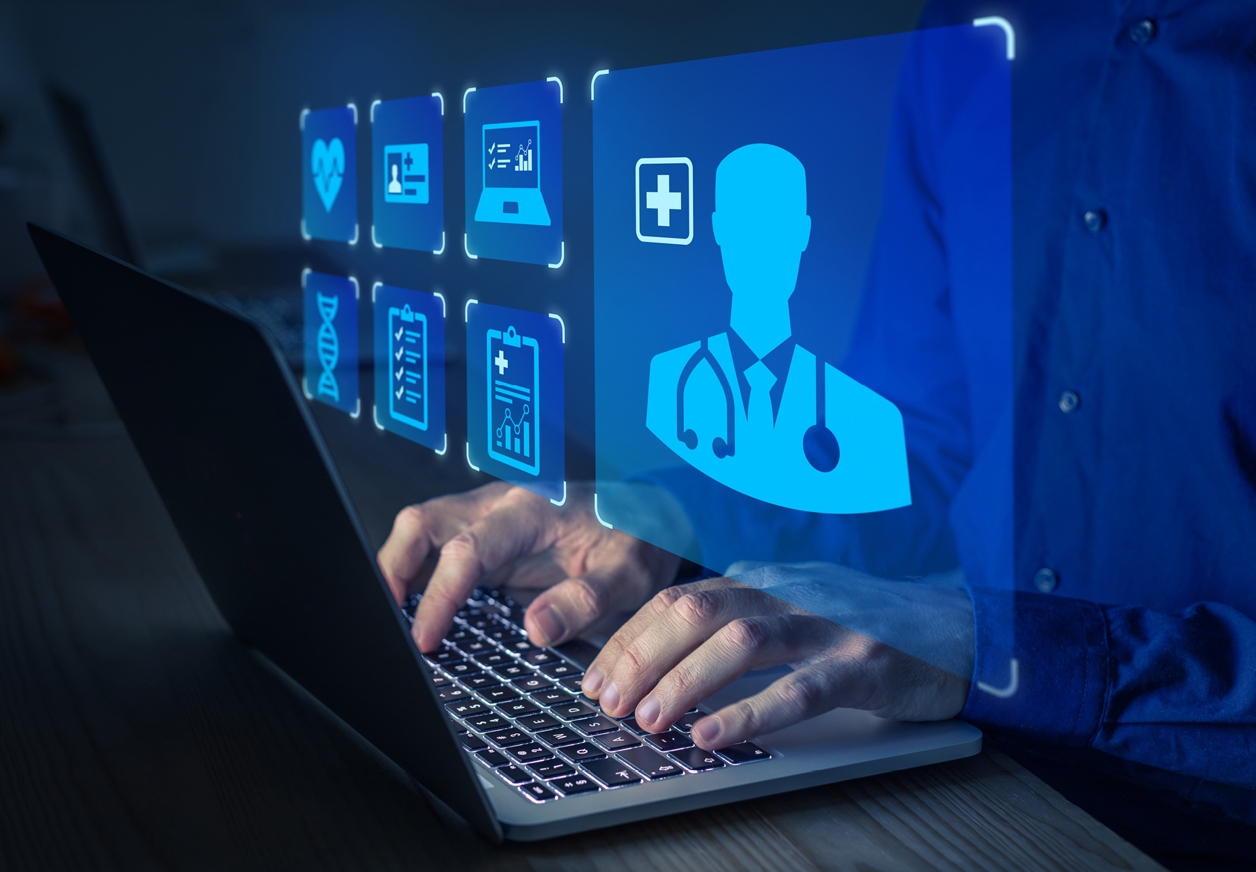 Closeup of someone typing on a laptop with blue medical-related graphic hovering over their hands. Dark background. Healthcare fraud concept. iStock image.