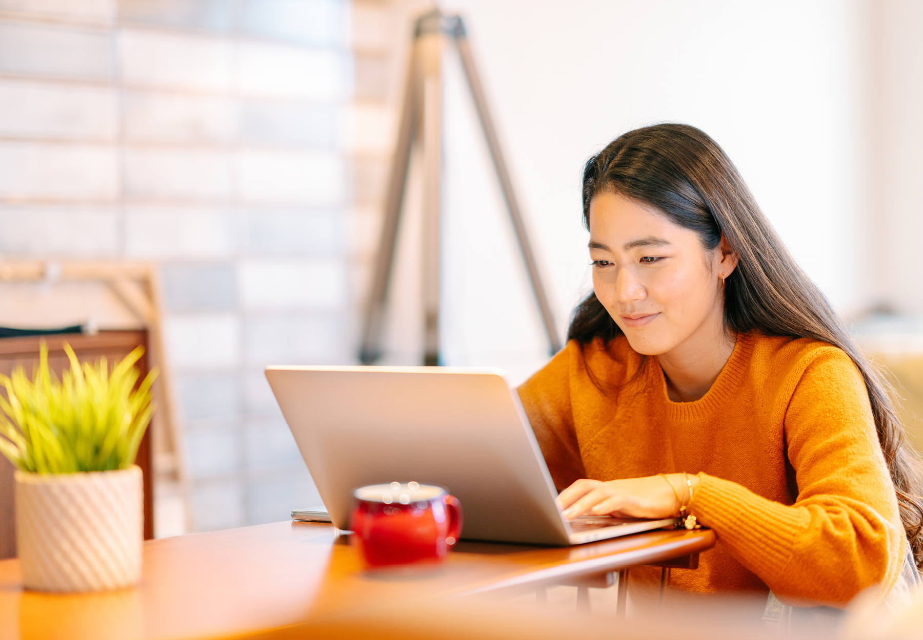 Woman in a dark yellow/light orange sweater is working on a laptop at a table in her home. Remote work concept. iStock image.