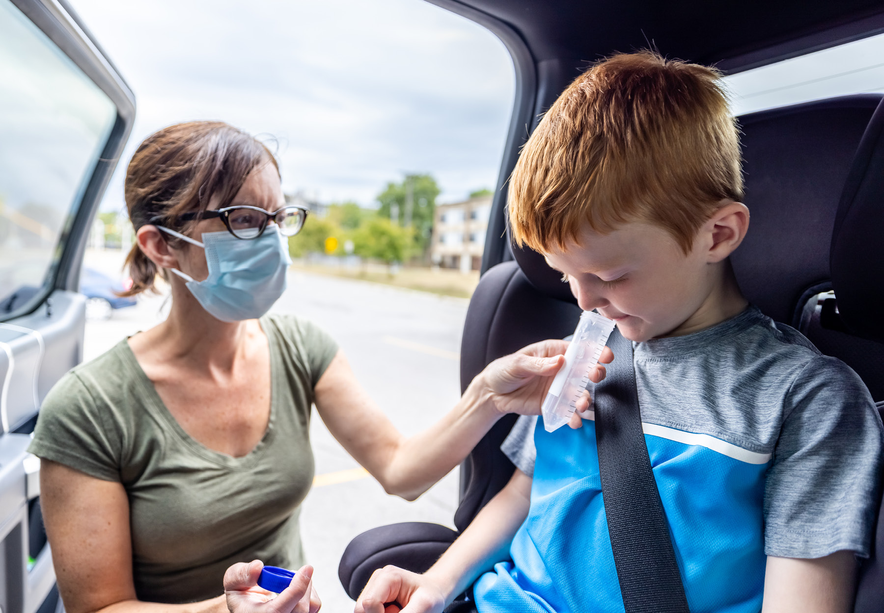 Mother in a mask is getting a saliva sample from her son for COVID-19 testing who is sitting in a car seat in a vehicle. iStock image.