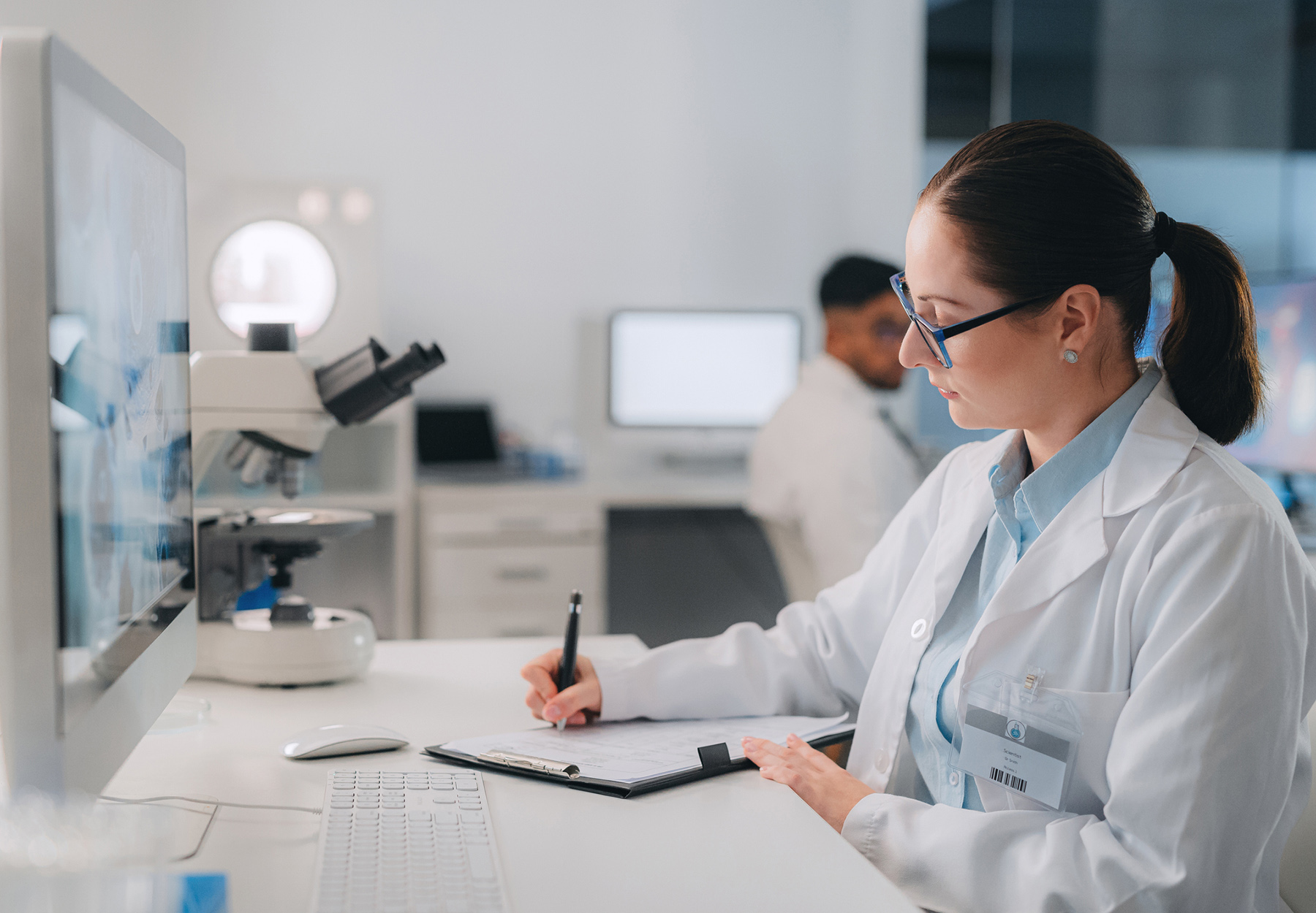 A female laboratory professional with glasses and a labcoat is sitting at a desk and writing notes on a clipboard. iStock image.